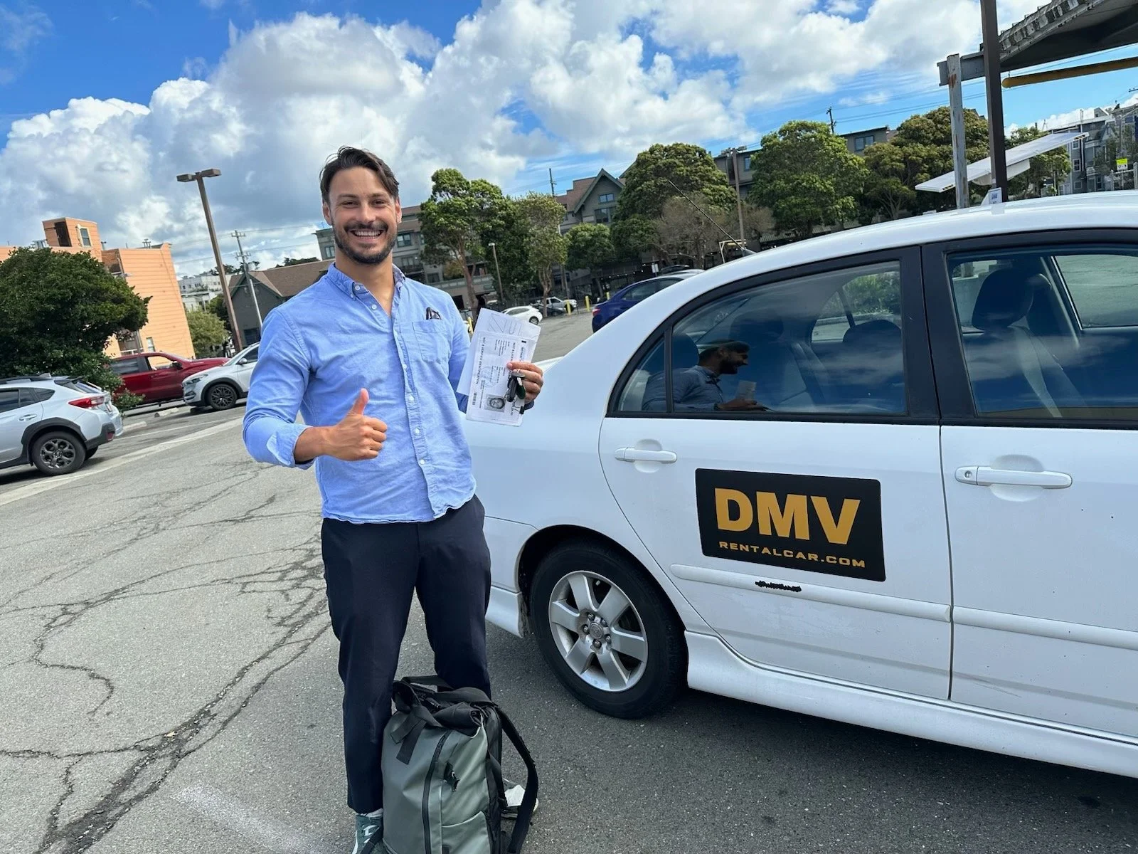 A man with dark hair and a beard, wearing a light blue shirt and dark pants, stands next to a white DMV rental car, giving a thumbs up and holding documents. There is a backpack on the ground in front of him. The background shows a parking lot with several cars, trees, and a partly cloudy sky.