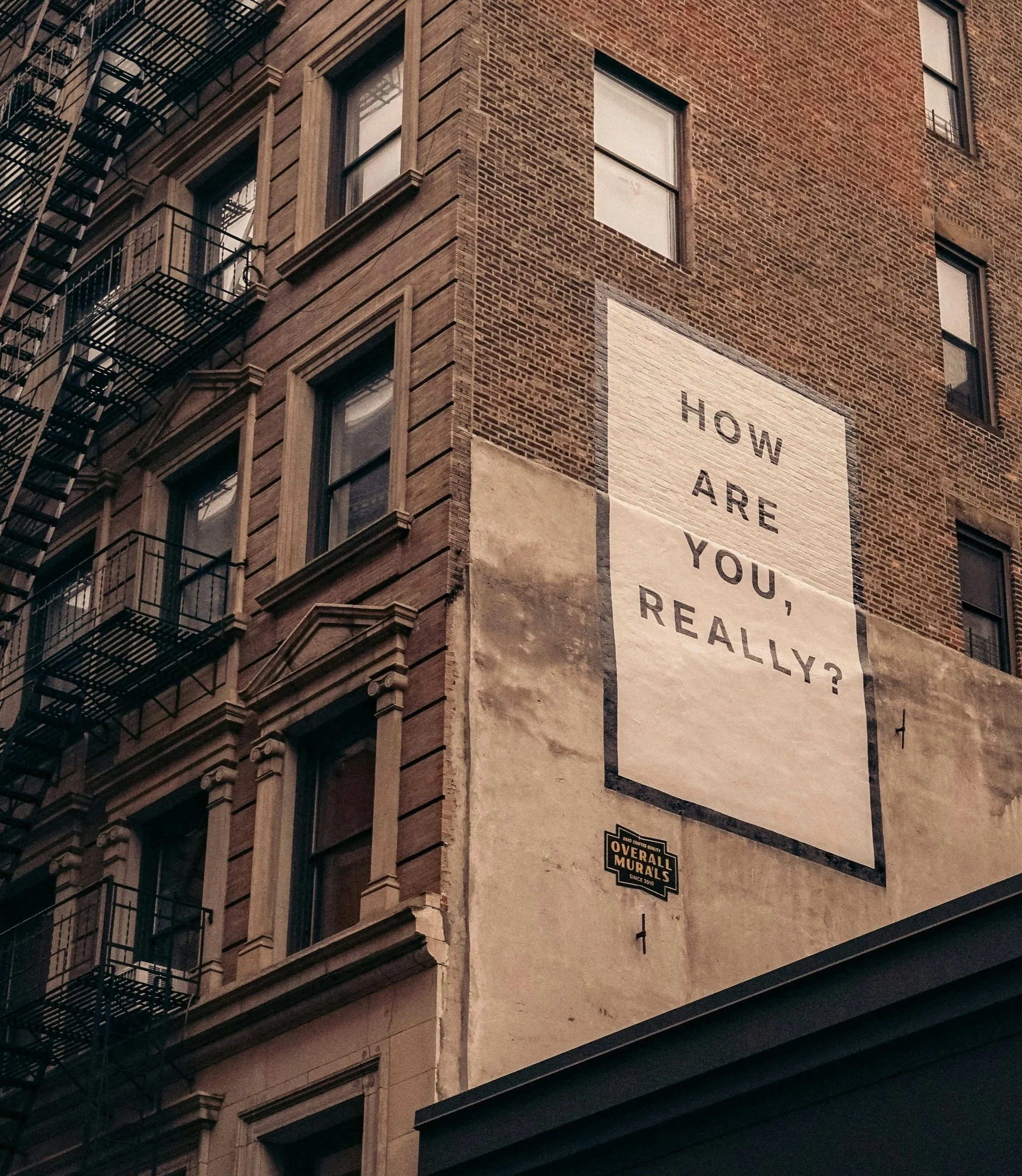 Brick city building with a large painted mural on the wall that reads "How are you, really?" Adjacent to windows and fire escapes.