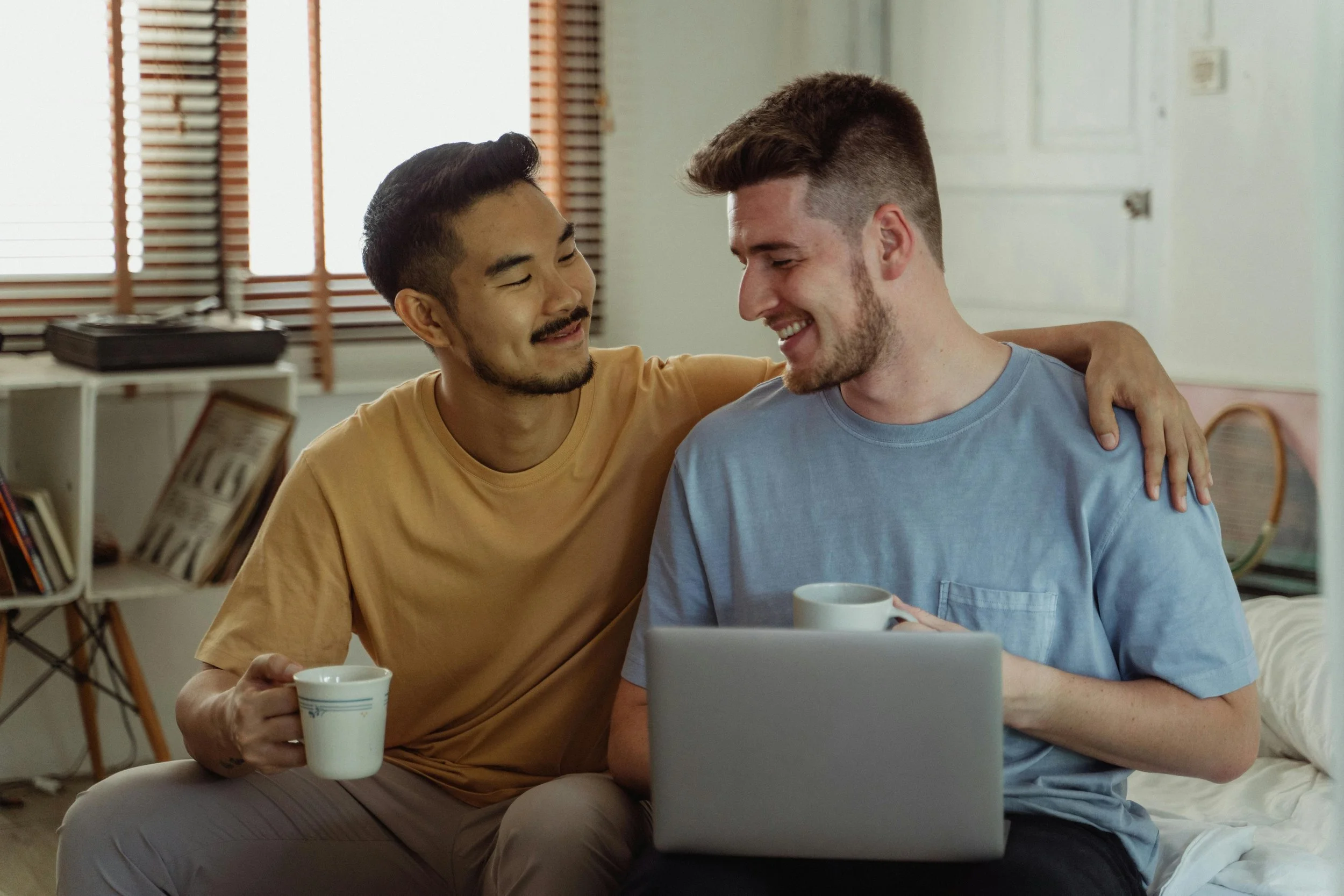 Two men sitting together, smiling and holding coffee mugs, with a laptop in front of them.