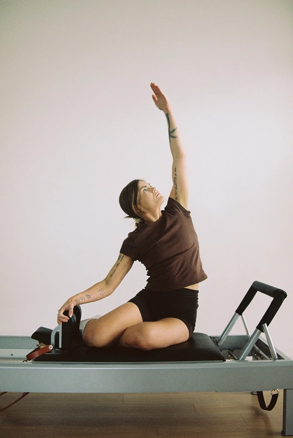 A woman practicing yoga on a reformer machine, sitting on her knees, reaching upward with her right arm and holding onto the reformer's handle with her left hand, against a plain wall.