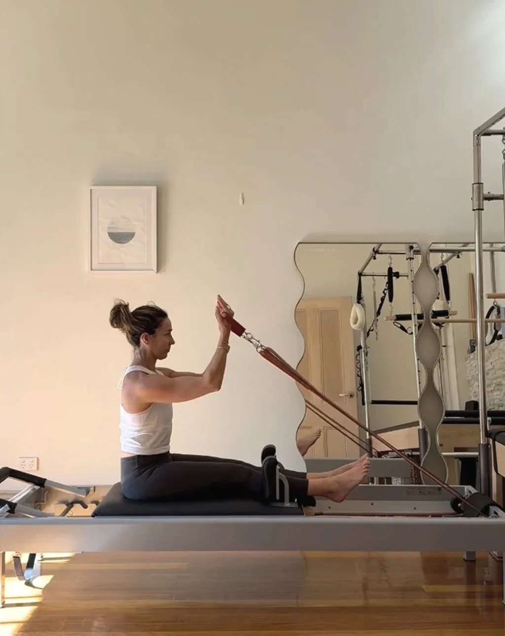 A woman exercising on a reformer pilates machine, pulling on resistance straps while sitting upright with her legs extended on the padded surface.