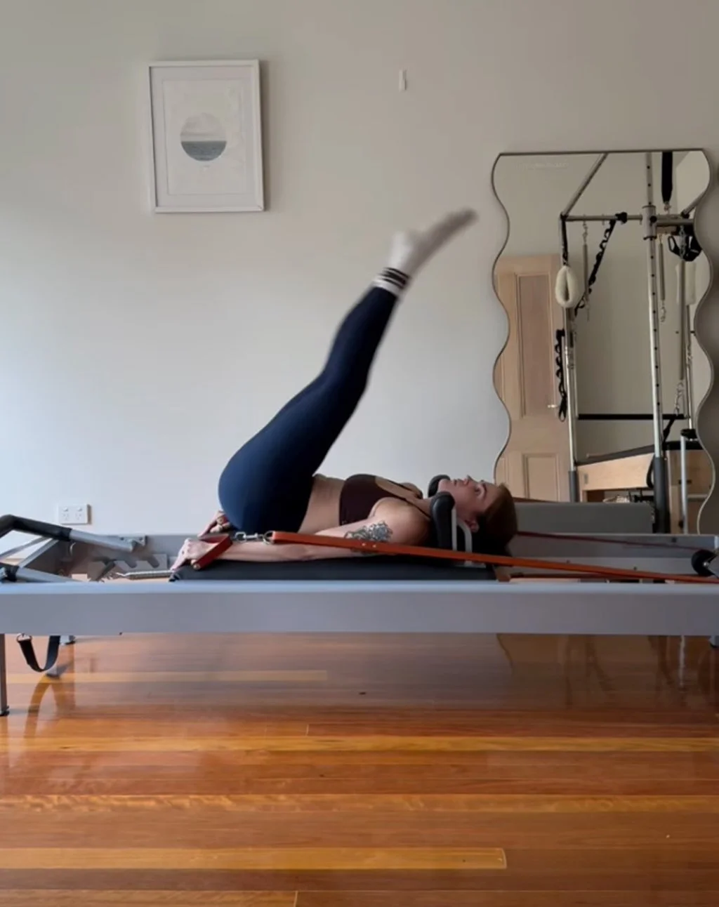 A woman lying on a Pilates reformer machine performing an exercise with one leg extended upward. She is in a fitness studio with wooden flooring, a large mirror, and gym equipment in the background.