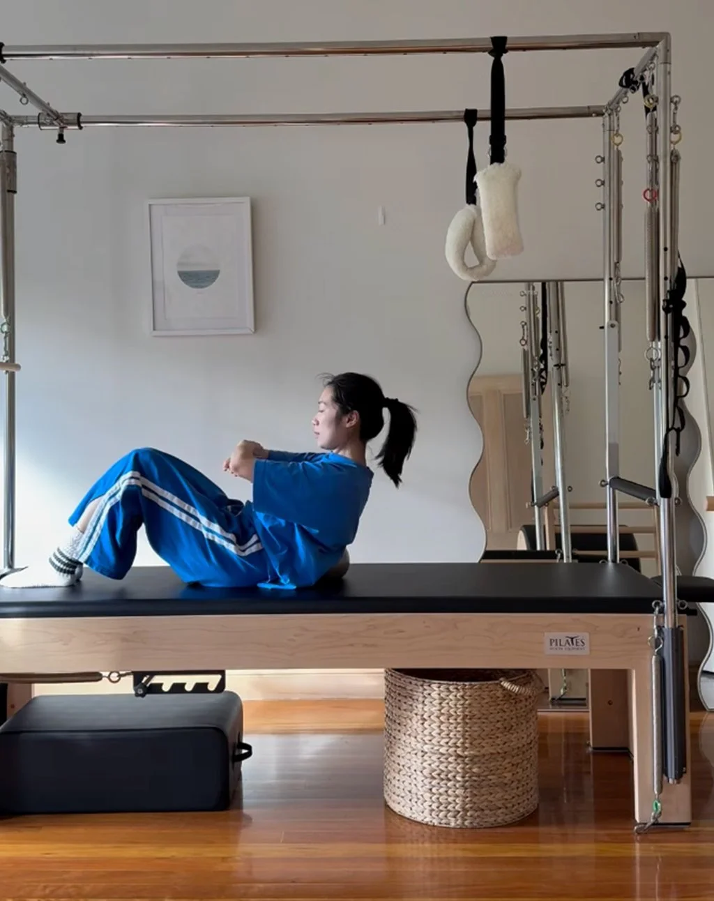 A woman in blue athletic clothing sitting on a Pilates reformer machine in a home studio, with a white wall, wall art, and exercise equipment around her.