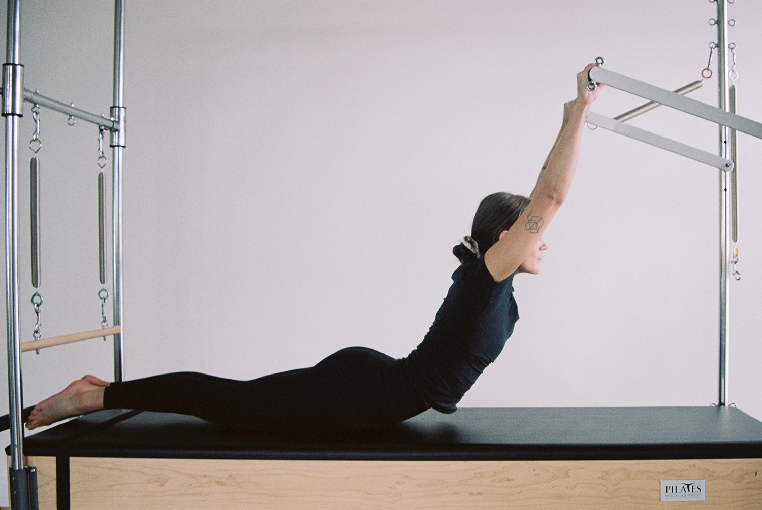 Woman practicing Pilates on a Reformer machine, lying prone with arms extended overhead, in a fitness studio.