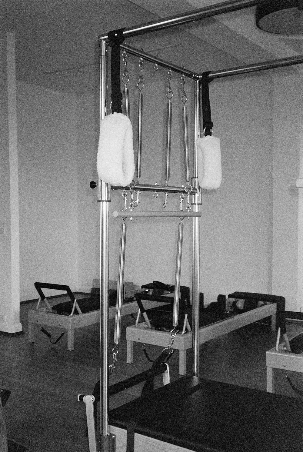 Reformer Pilates machine with adjustable straps and rectangular pads, in a fitness studio with additional reformer equipment in the background.