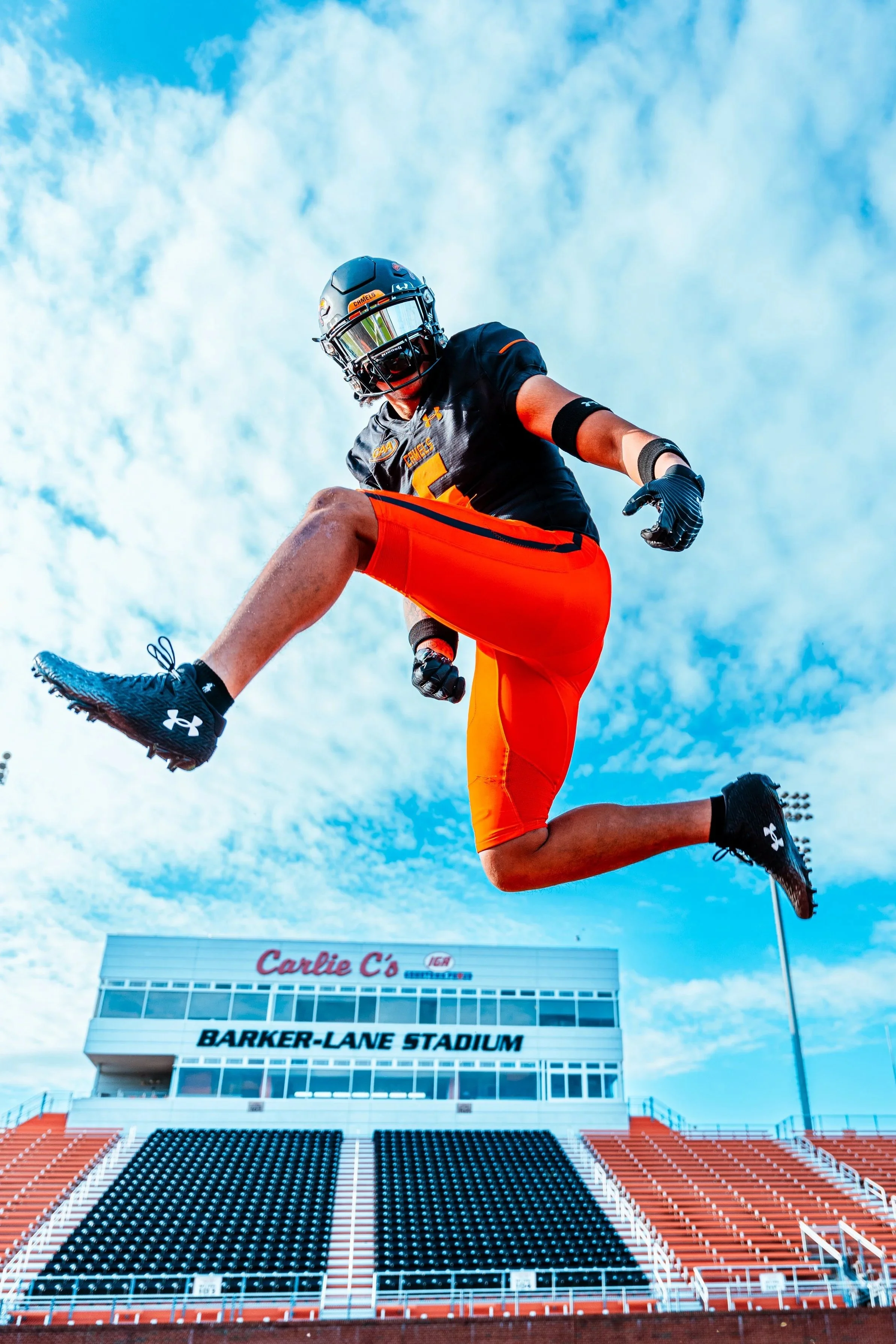 An athlete in a football uniform jumps in the air with the football in hand at Barker-Lane Stadium, with blue sky and clouds above.
