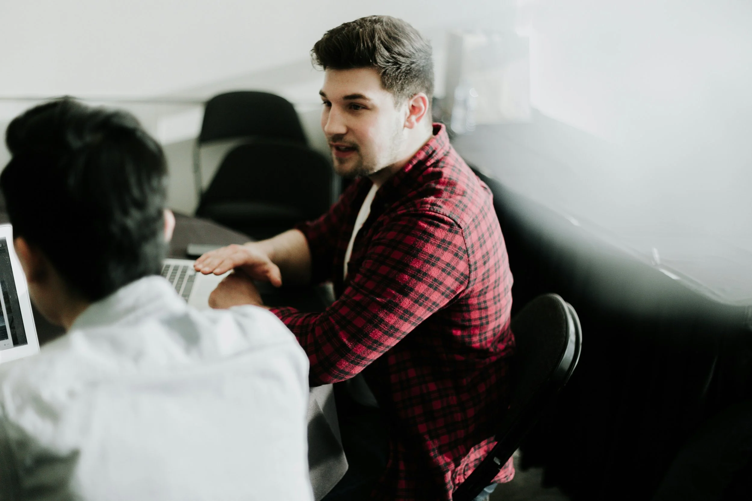 Two people sitting at a table with laptops, engaged in conversation; one wears a red plaid shirt.