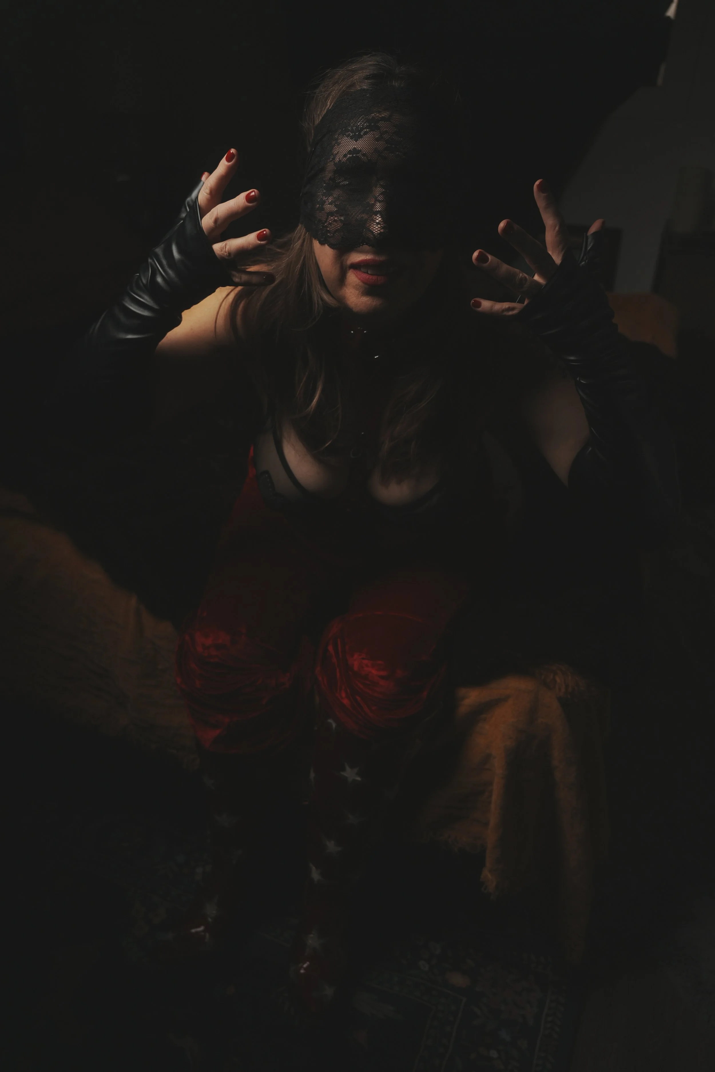 A woman dressed in dark clothing with a black lace blindfold covering her eyes, wearing black gloves with red nails, and sitting on a patterned rug in a dimly lit room.