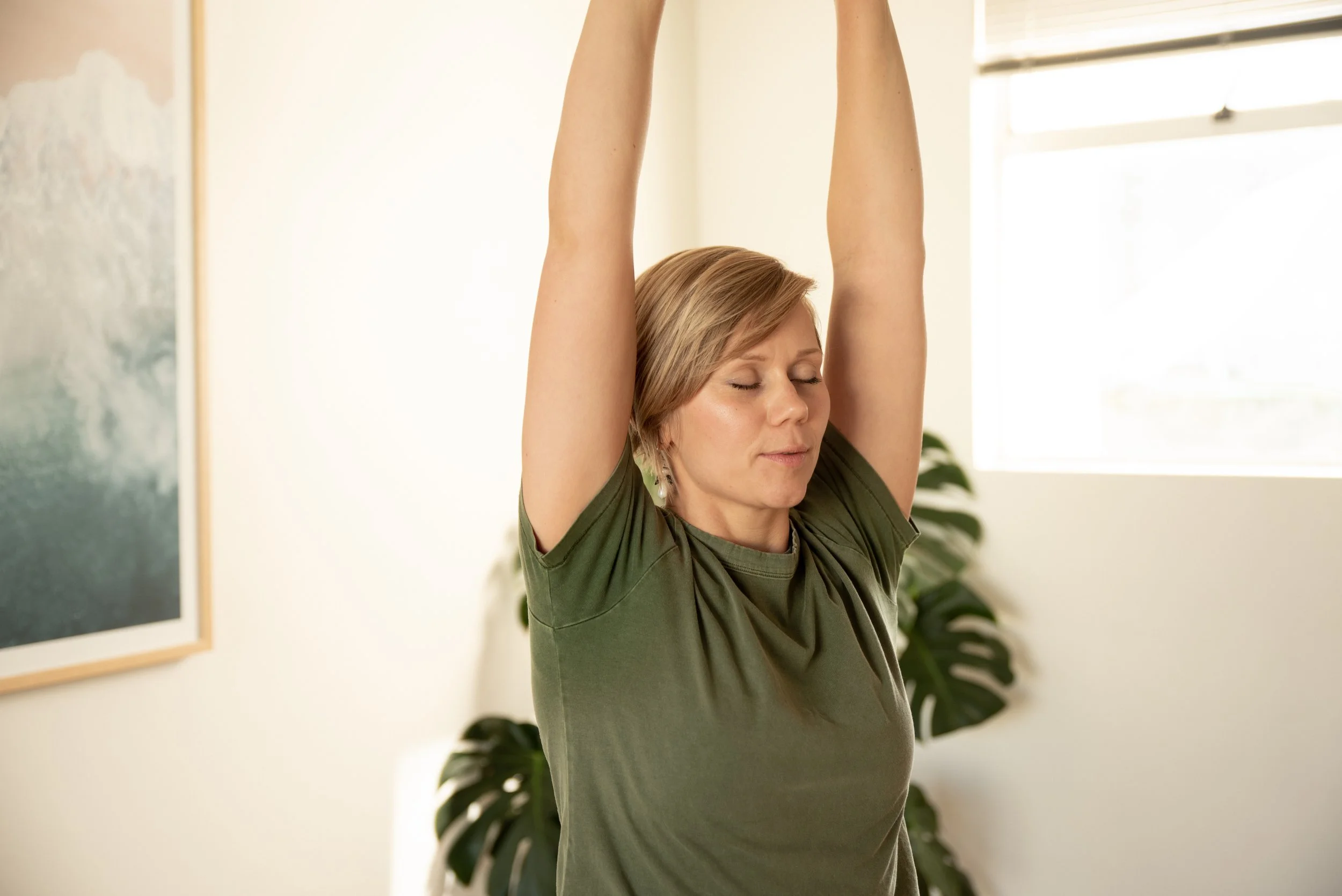 Vihreään paitaan pukeutunut henkilö venyttelee kädet kohotettuna valoisassa huoneessa.