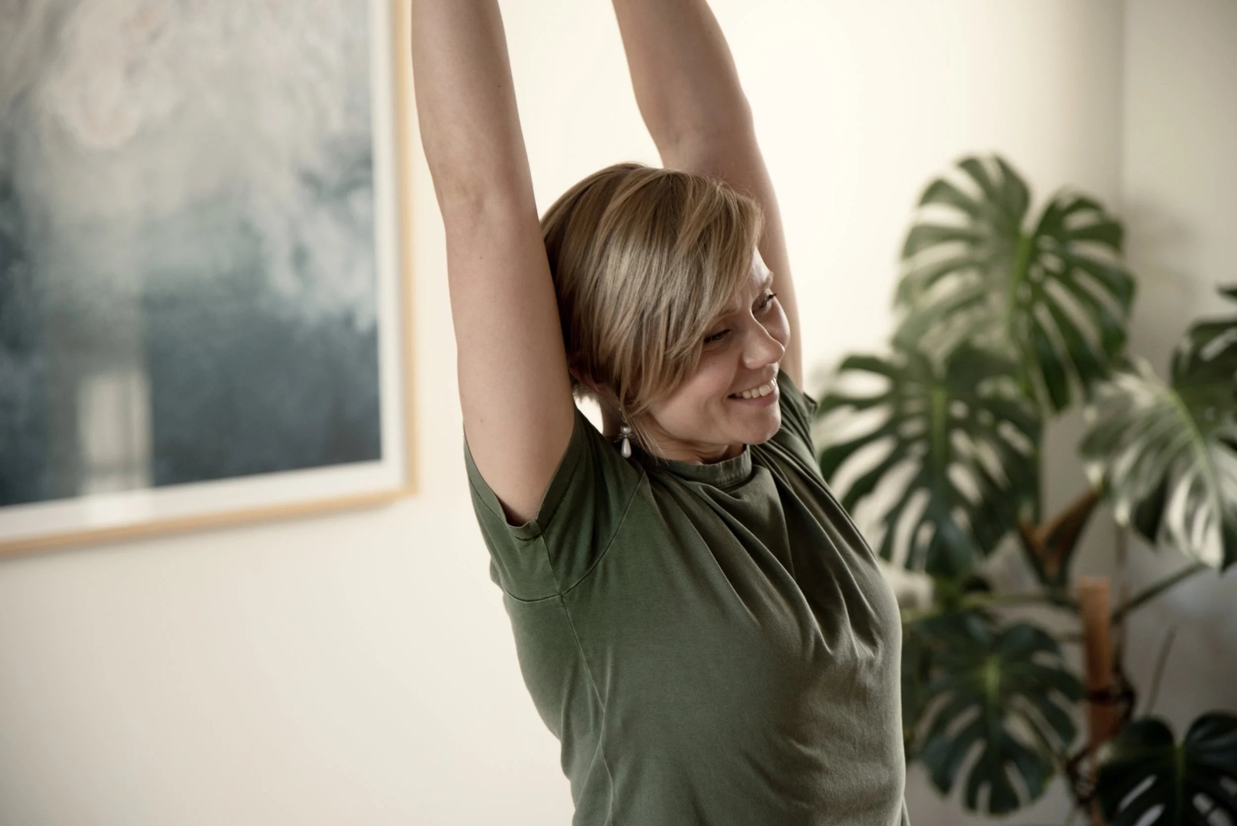 Vihreäpaitainen henkilö venyttelee kädet kohotettuna, hymyillen, suuren huonekasvin lähellä.