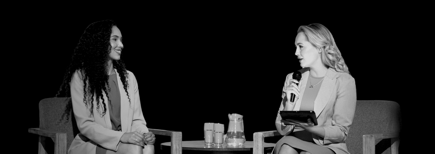 Two people seated in armchairs for an on-stage interview, one holding a microphone.