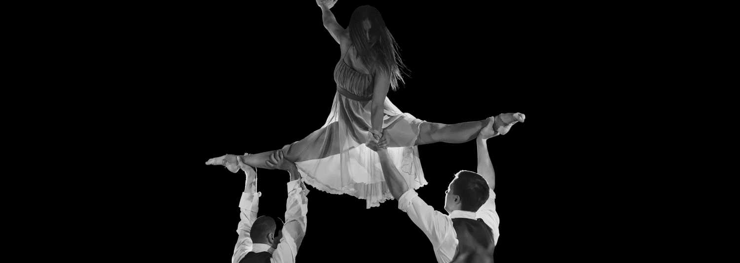 Three ballet dancers performing a lift, one dancer held in mid-air, against a black background.