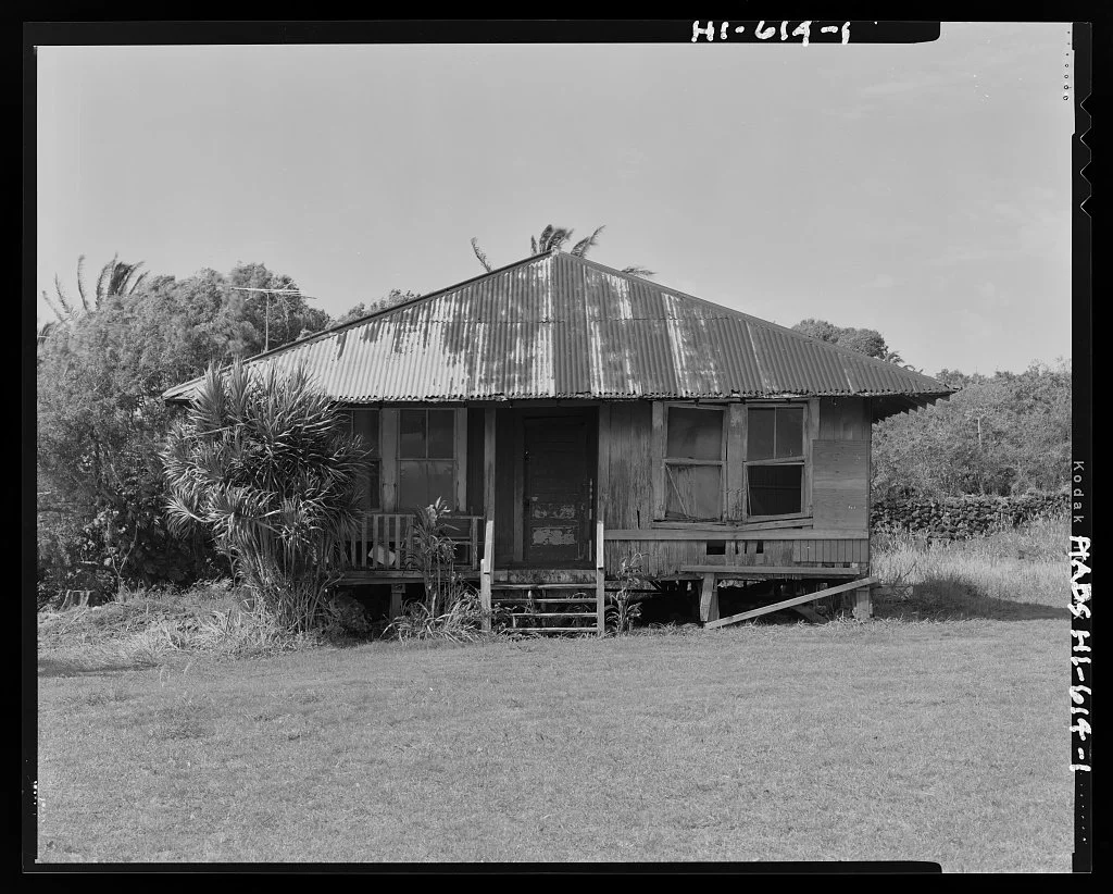 This black and white photograph shows the exterior of the Kaupo School Teacher's Cottage. The small, modest structure features a corrugated metal hip roof with visible weathering and a simple wooden construction elevated on posts.