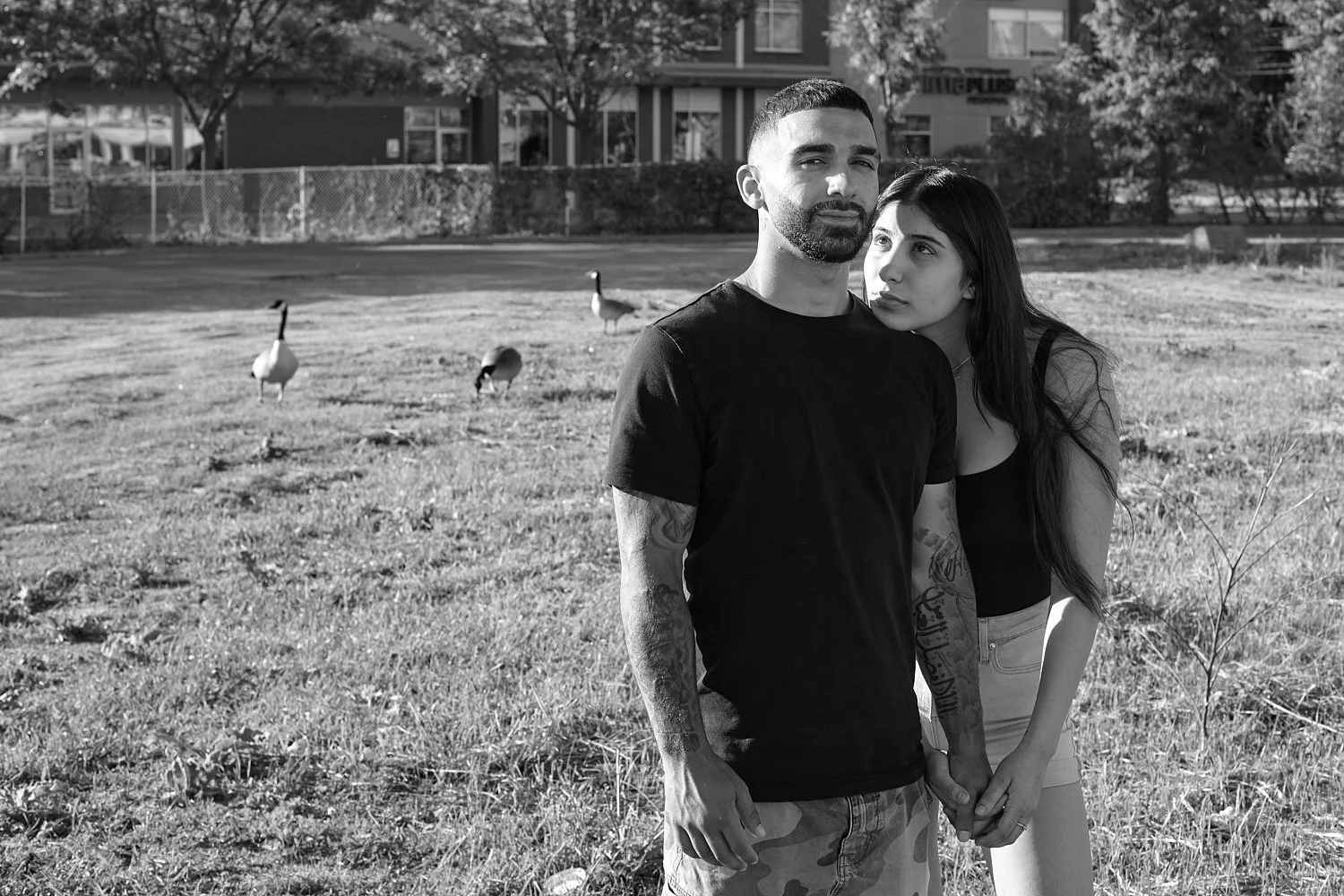 A black and white photo of a man and woman holding hands in a park with three geese in the background, behind a fence and trees.