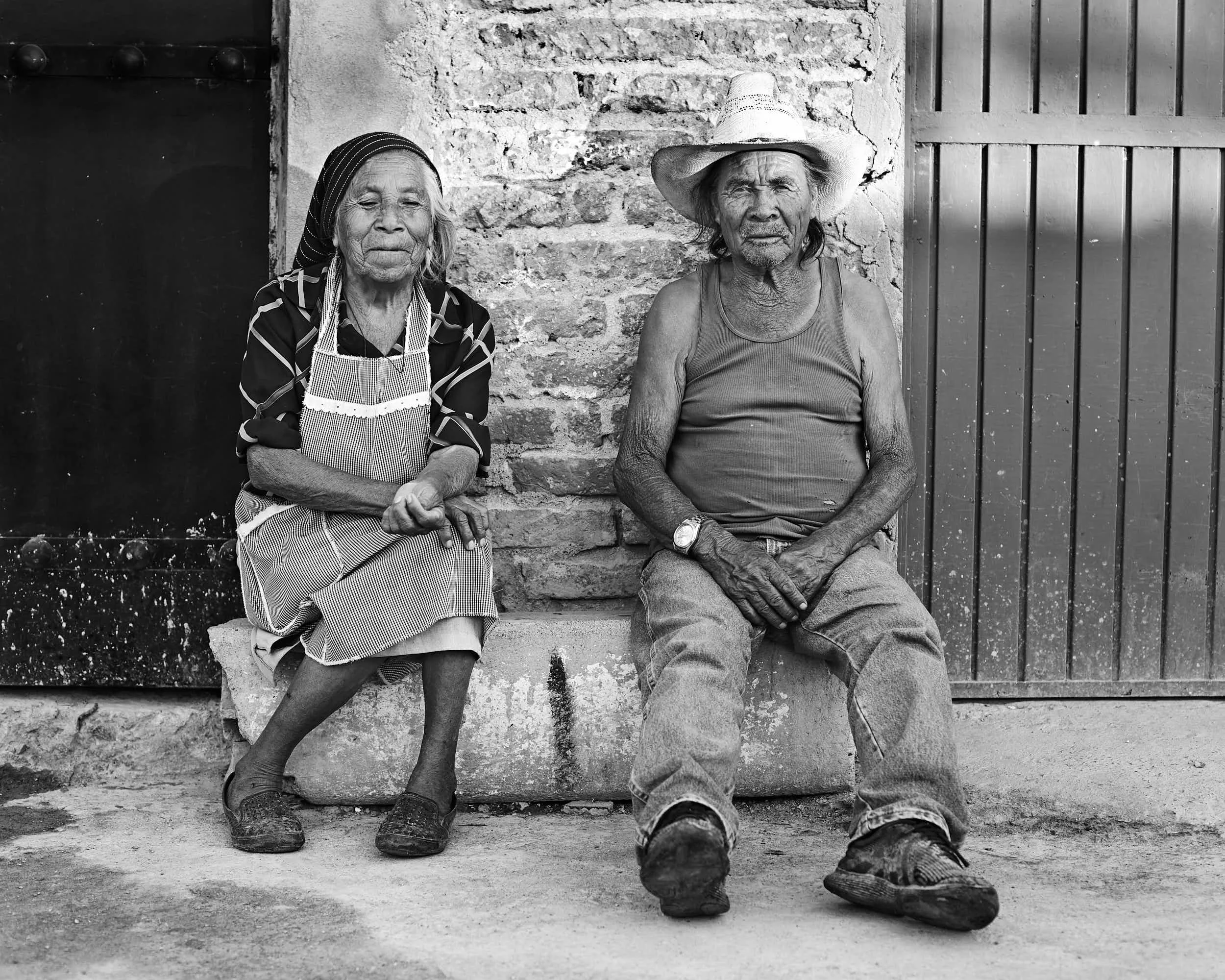 An elderly woman and man sitting on a stone step in front of a brick wall. The woman is wearing a headscarf, patterned dress, apron, and slippers. The man is wearing a straw hat, tank top, jeans, and sneakers, with a watch on his left wrist.