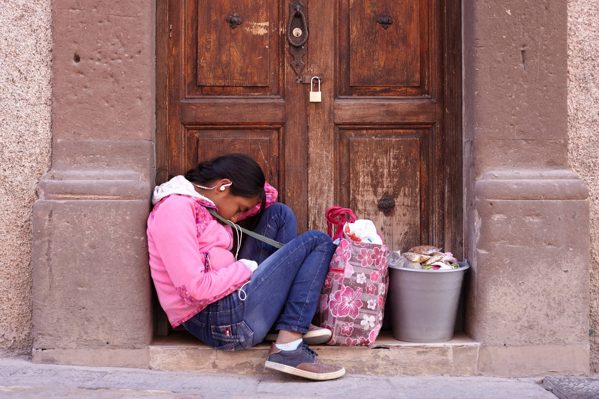 A girl slumped and sleeping on the ground against a wooden door, with a bag and a bucket filled with clothes beside her.