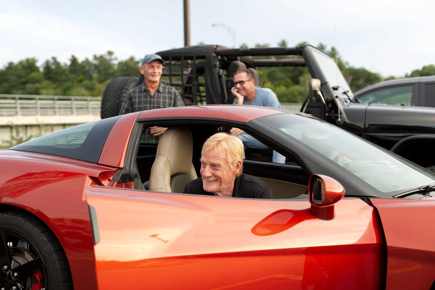 This image shows three men, one sharply in focus in the foreground inside a bright red orange sports car and laughing. The two men in the background are smiling and somewhat out of focus. Also visible behind the three of them is a bridge and an SUV v