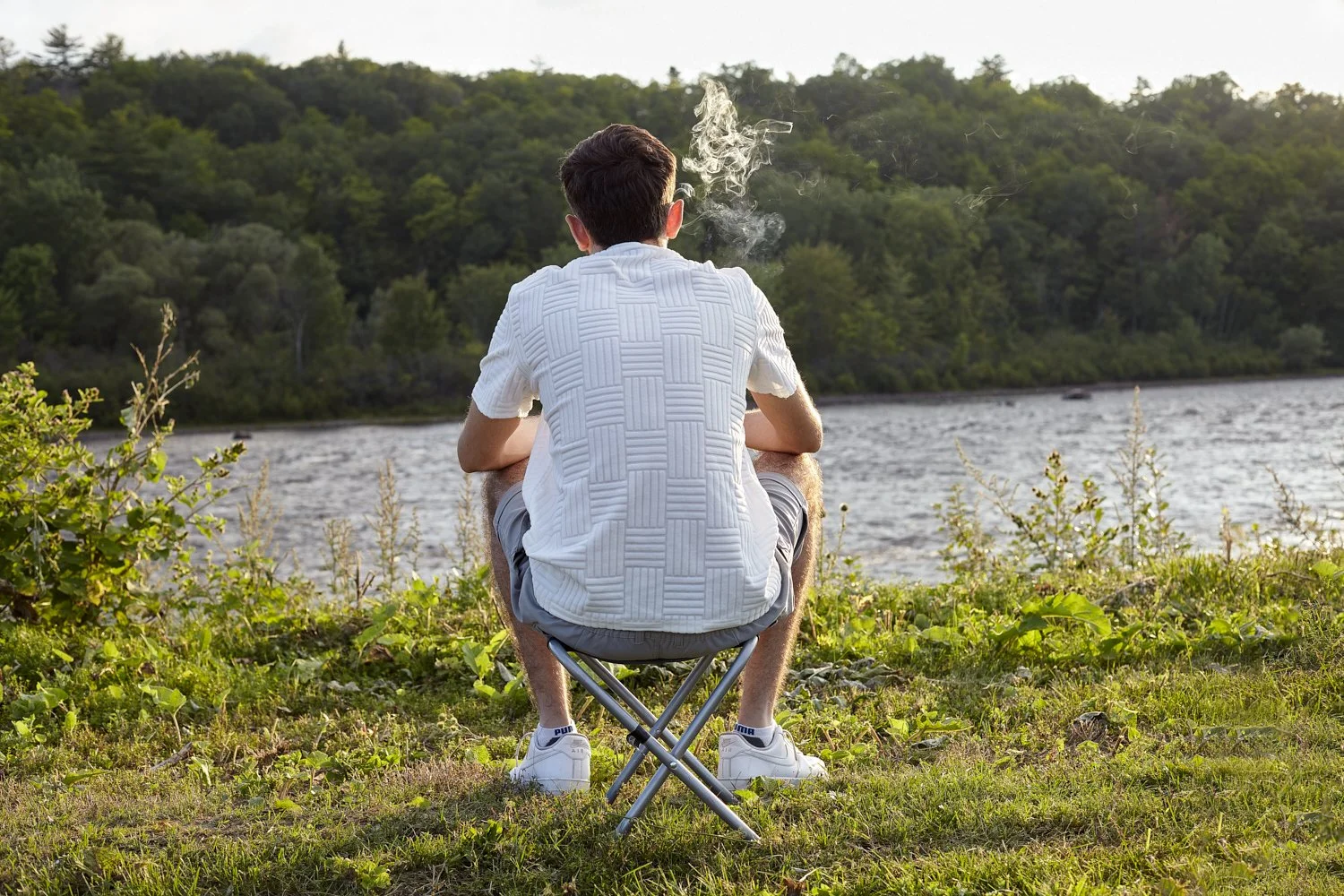 A young man with short hair, wearing a short-sleeved shirt, shorts, and sneakers, sits on a small portable stool near a body of water, facing away from the camera, with smoke rising from what appears to be a cigarette or vape in his hand. Trees and h