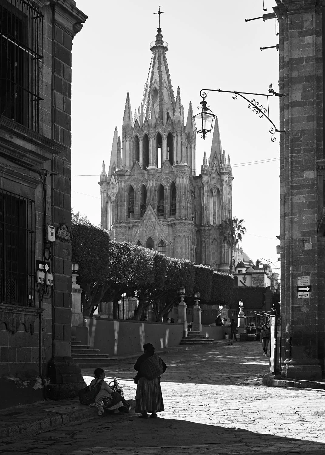 A black and white photo of a street scene with a large, ornate church in the background, and two women talking in the foreground, one sitting on the ground with a bag and the other standing. The church has multiple spires and a cross on top.