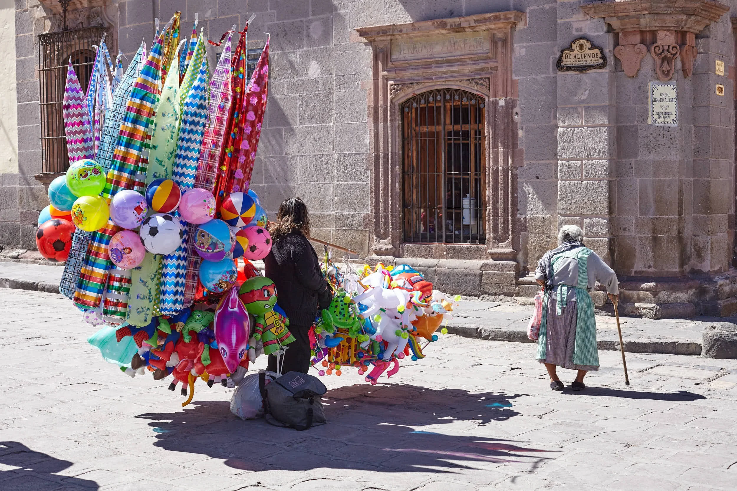 Street vendor sells colorful inflatable toys and balls, with an elderly woman walking past on a cobblestone street.