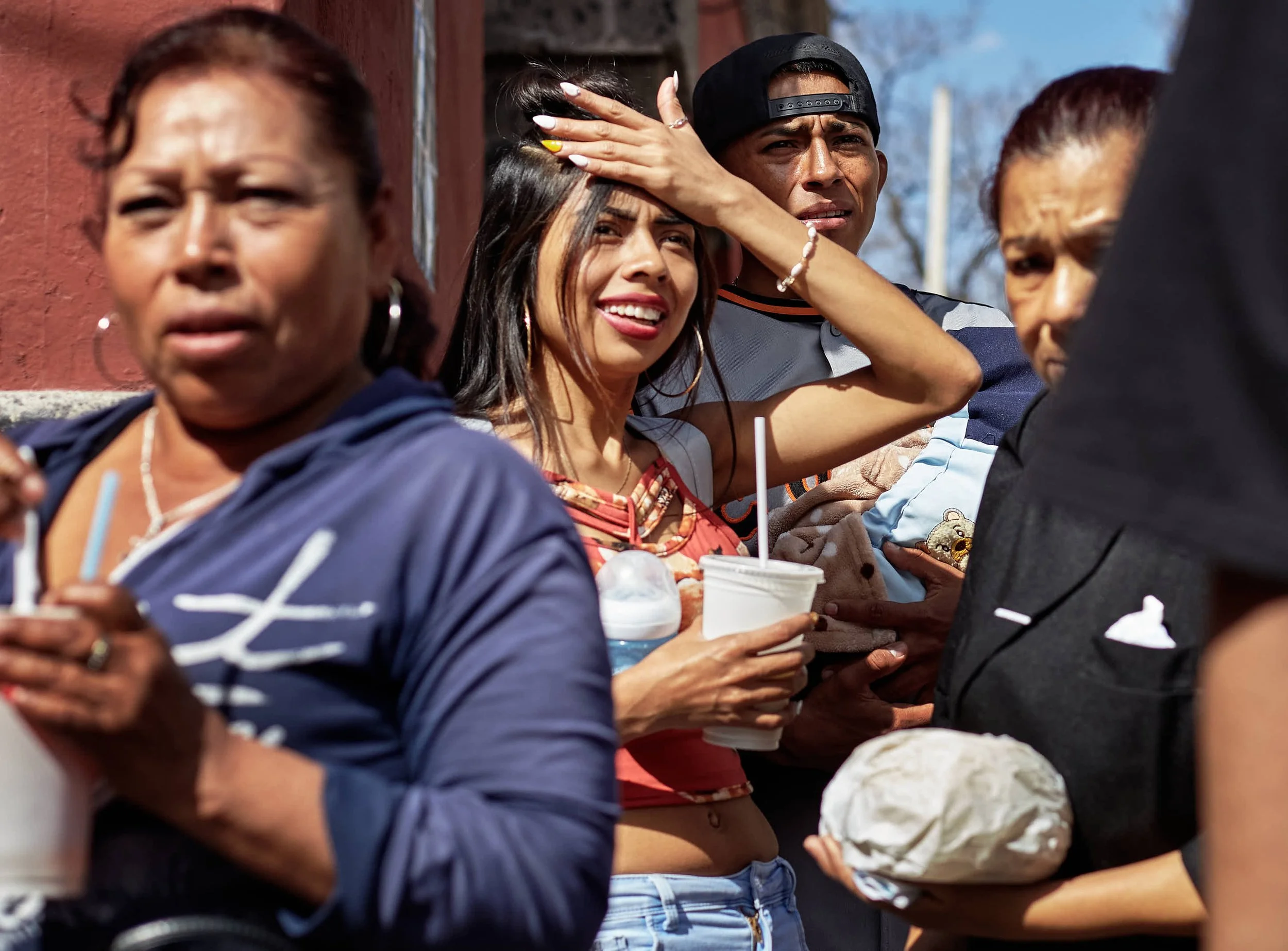 A group of people moving along a crowded sidewalk, some holding drinks and food, a woman shielding her eyes from the sun and a man behind her with a baby.