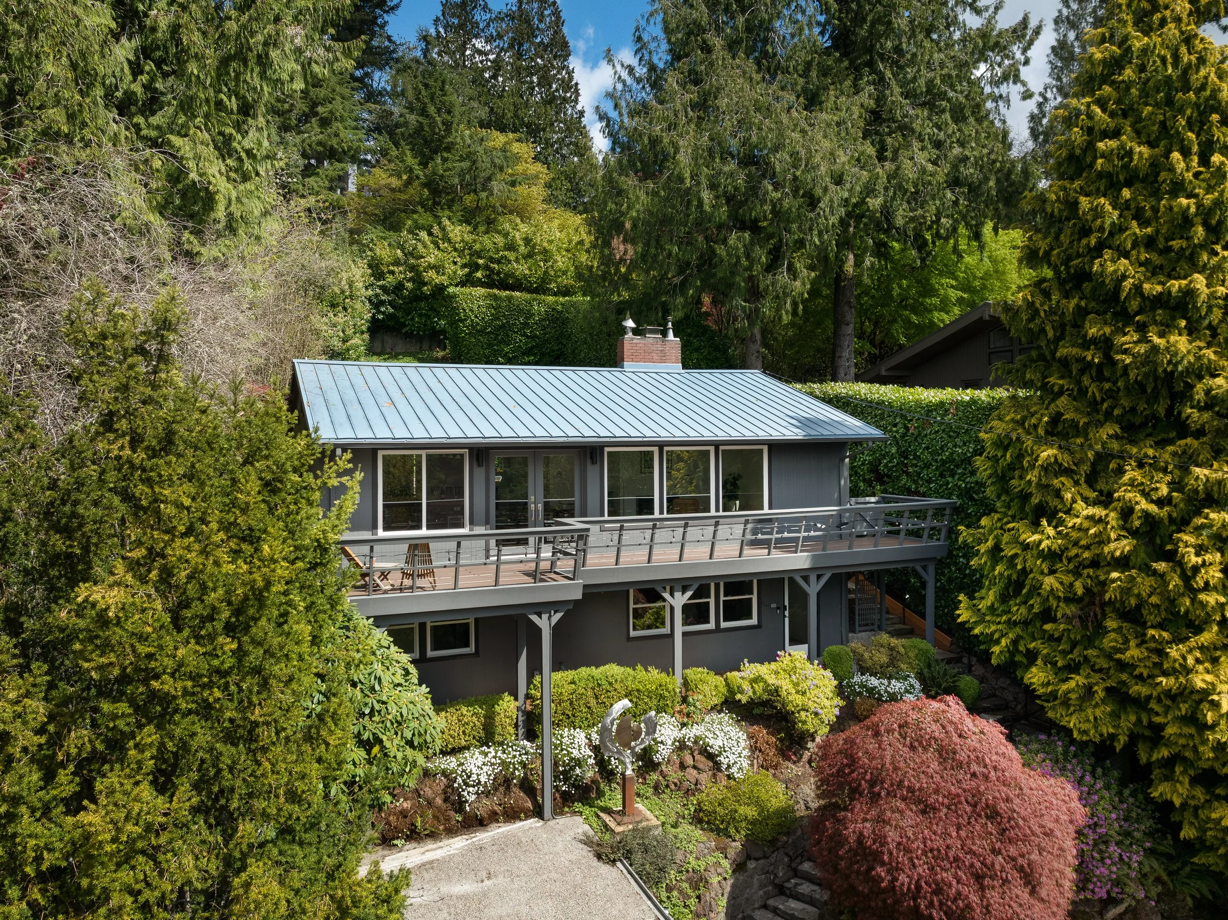 A house with a gray metal roof surrounded by lush green trees and colorful flower bushes in a garden.