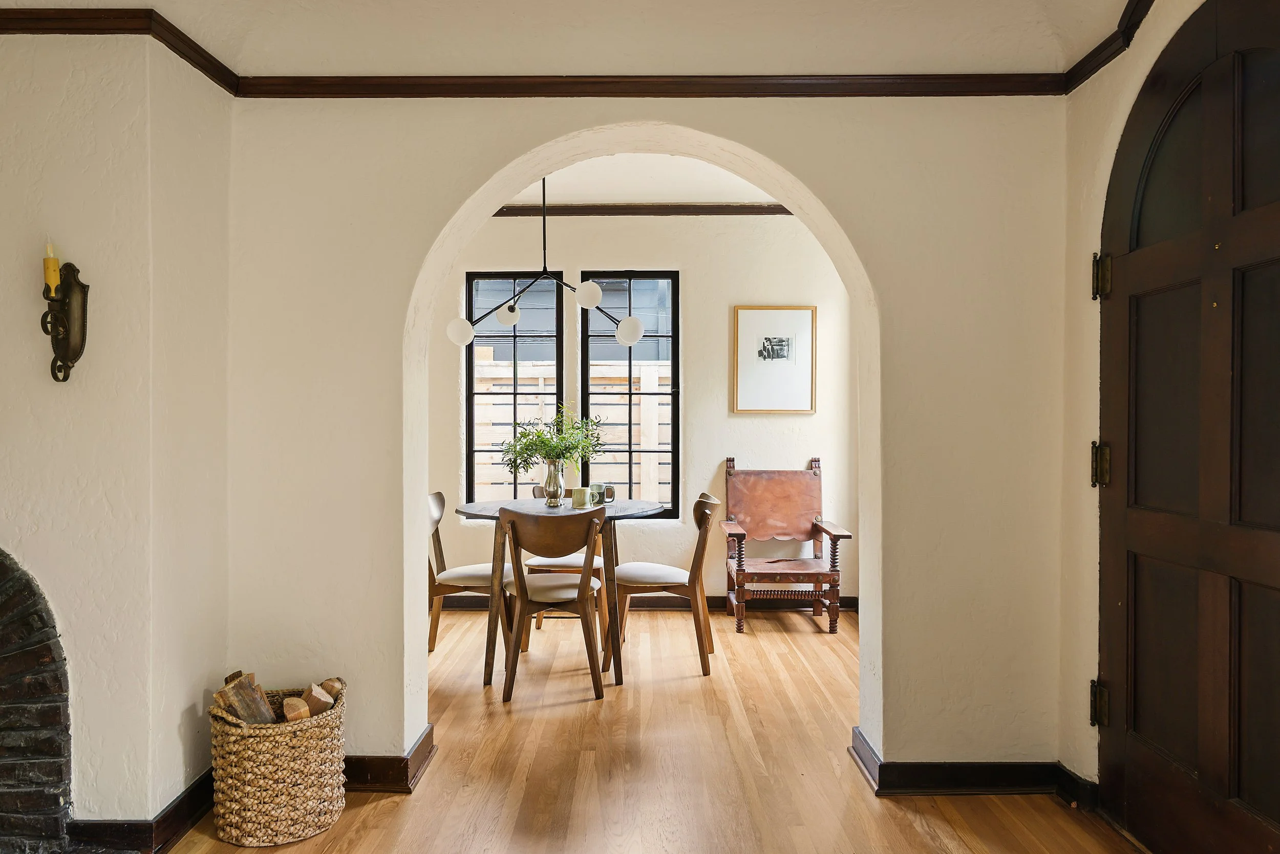 A cozy dining area with a round table, four wooden chairs, and a window with black grid panes. The room has light beige walls, a wooden floor, a modern chandelier hanging above the table, and framed artwork on the wall. There is a chair with a leathe