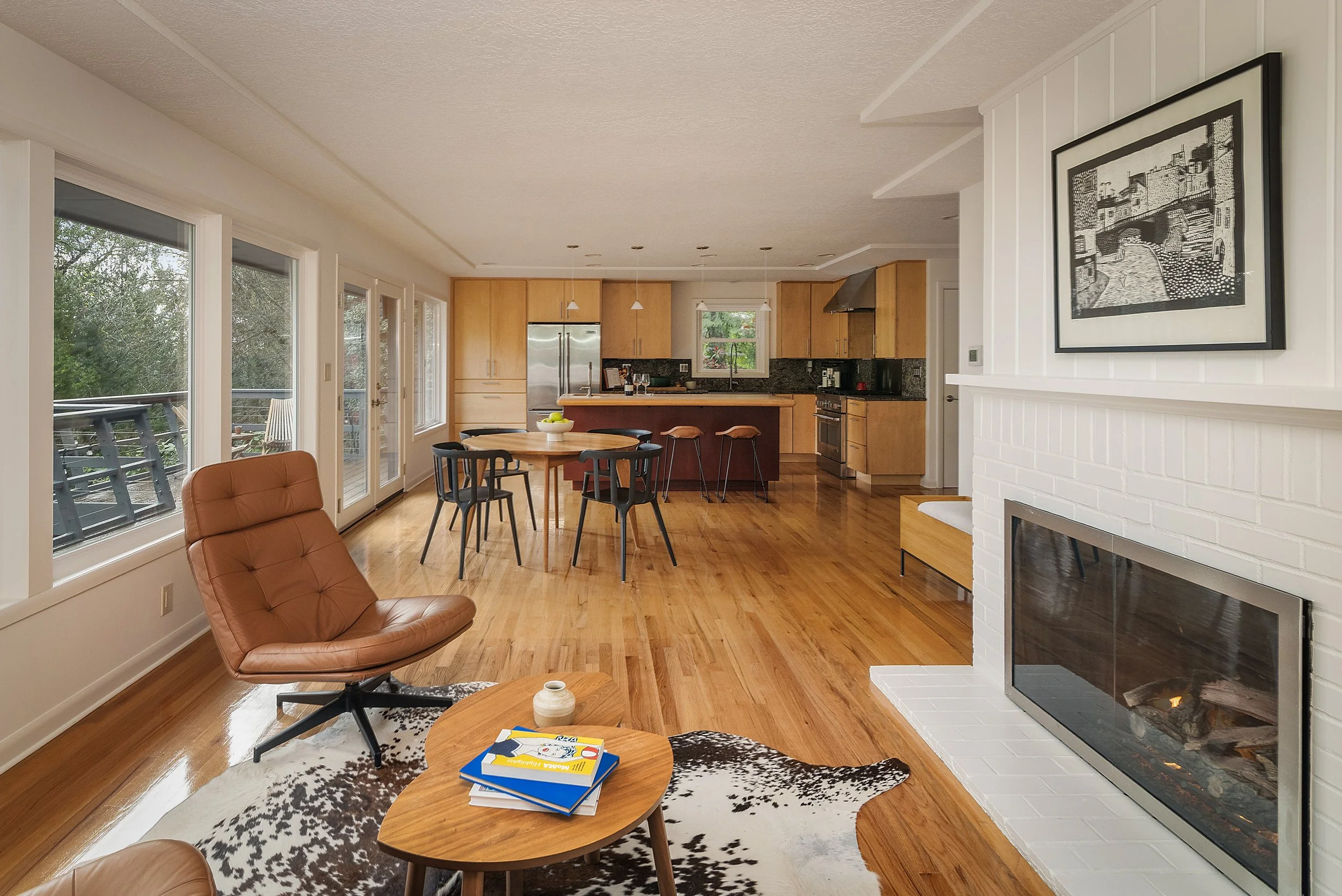 A cozy living room with hardwood floors, a brown leather swivel chair, a black and white cowhide area rug, and a wooden coffee table with books and a small decorative item. Large windows and a sliding glass door let in natural light. The kitchen in t