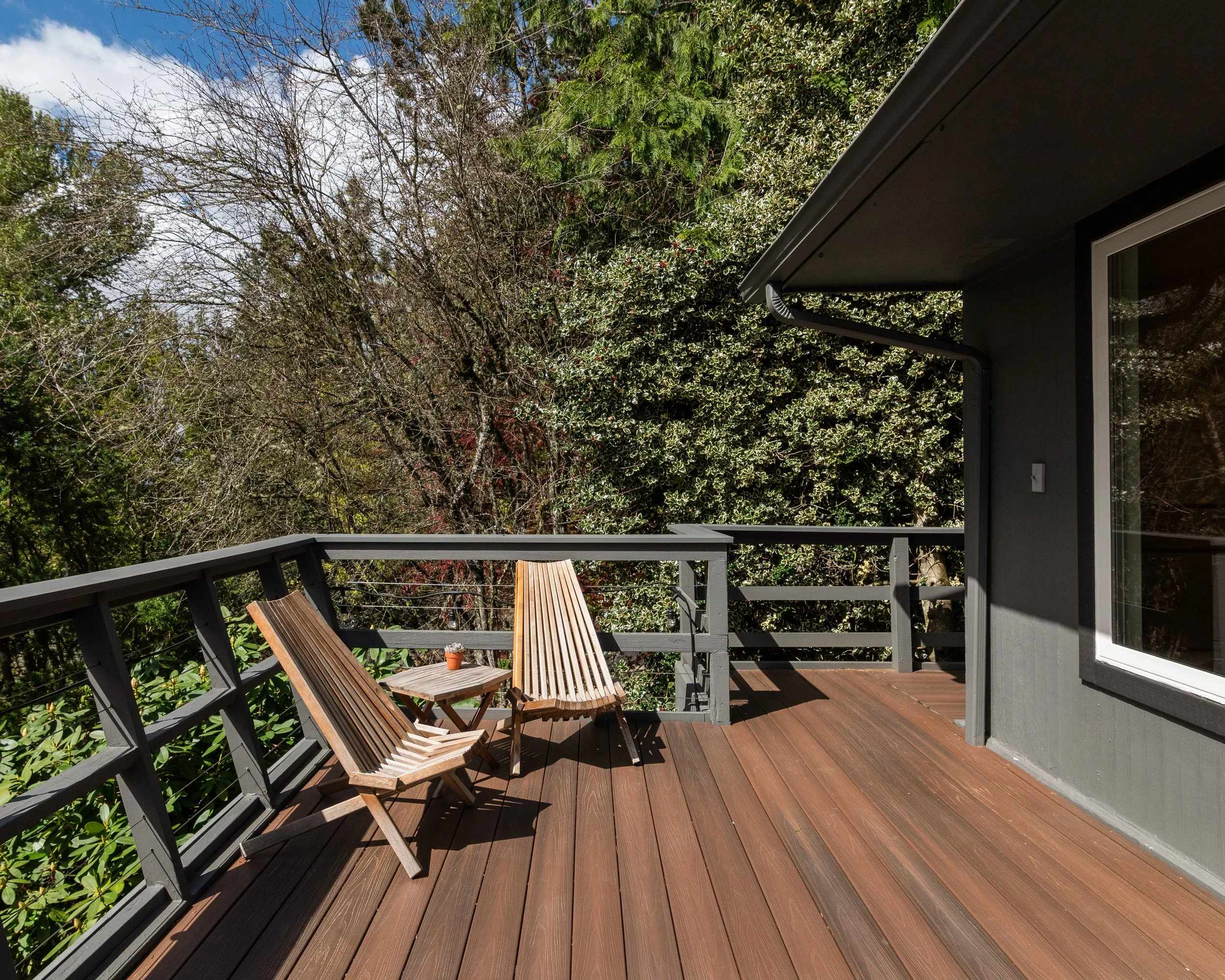 A house balcony with wooden chairs and a small table, overlooking green trees under a partly cloudy sky.