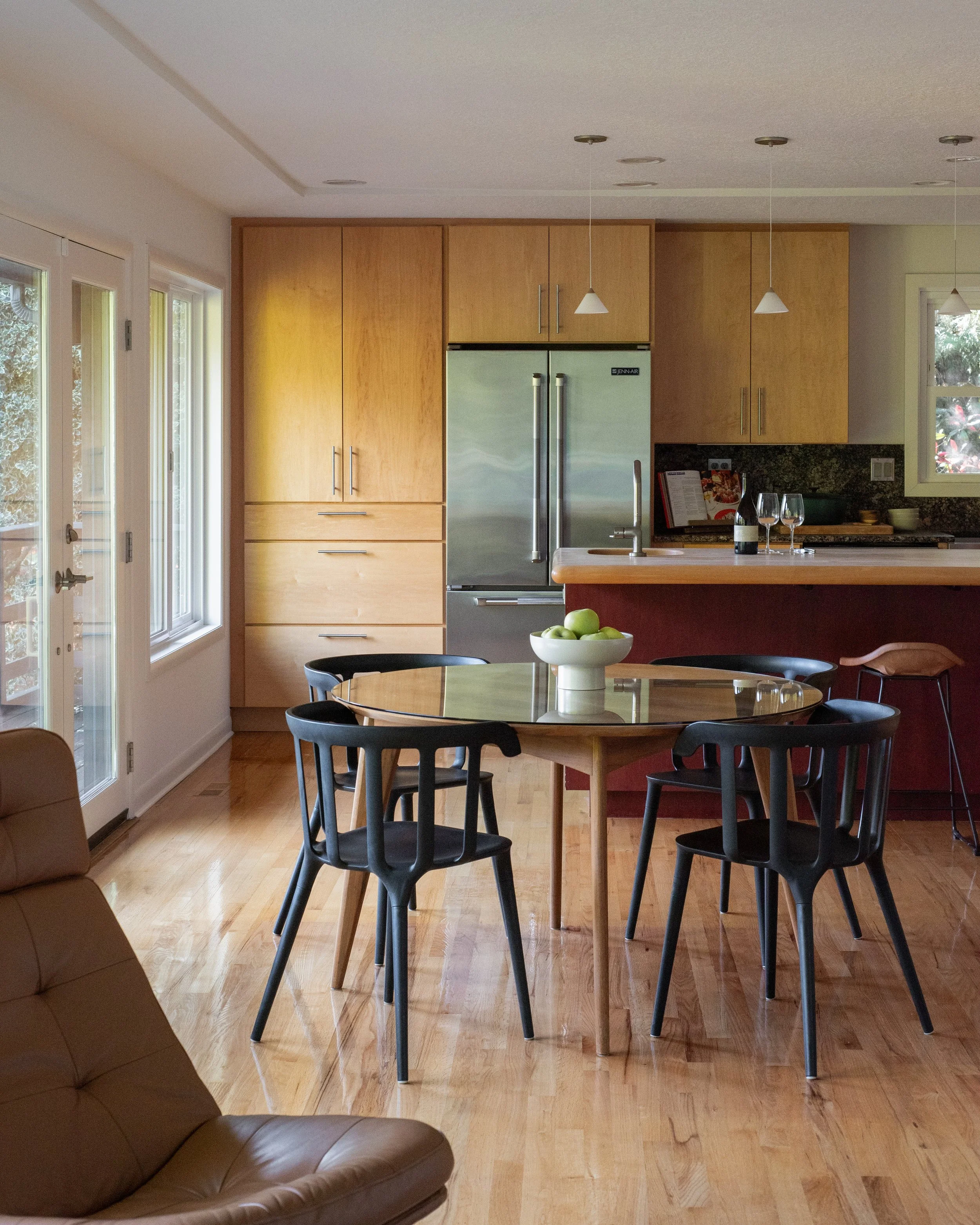 A modern kitchen and dining area with a round wooden table, black chairs, and a bowl of green apples. The kitchen has light wood cabinets, a stainless steel refrigerator, and pendant lights hanging from the ceiling. There are large windows on the lef