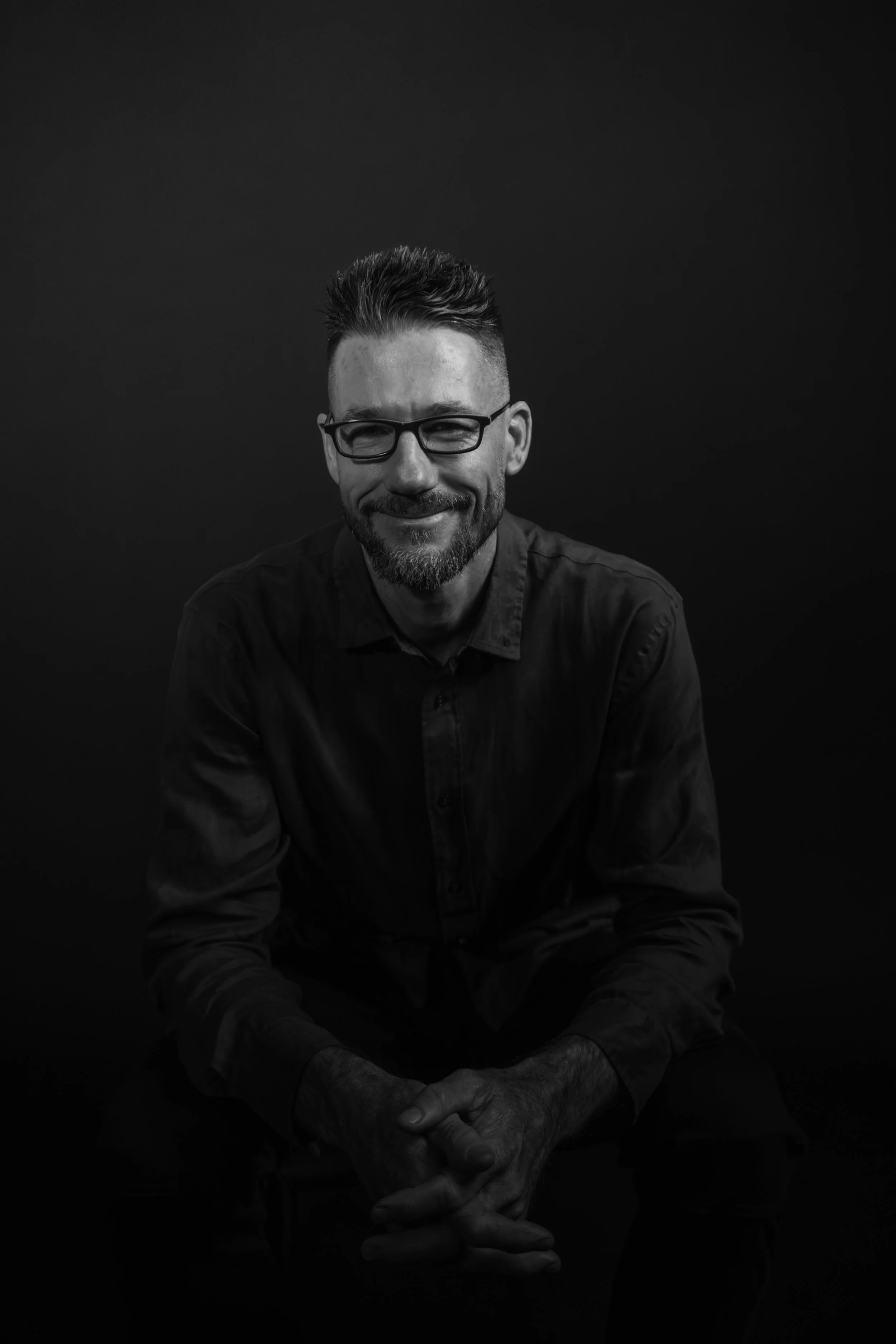 Black-and-white portrait of a middle-aged man with glasses, a beard, and styled hair, smiling softly, sitting against a dark background.