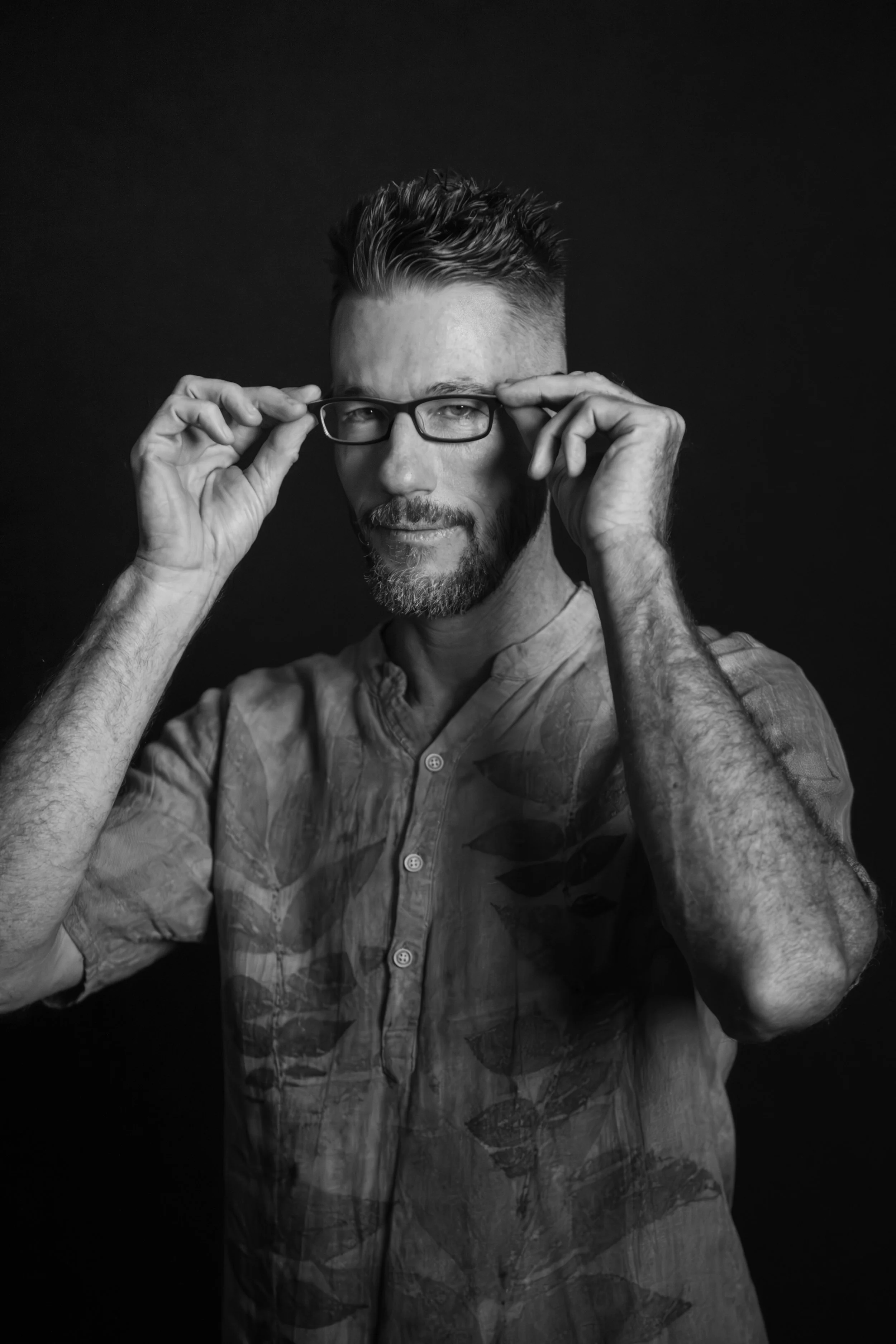 A black-and-white portrait of a man with a beard and glasses, looking at the camera, adjusting his glasses with both hands, against a dark background.