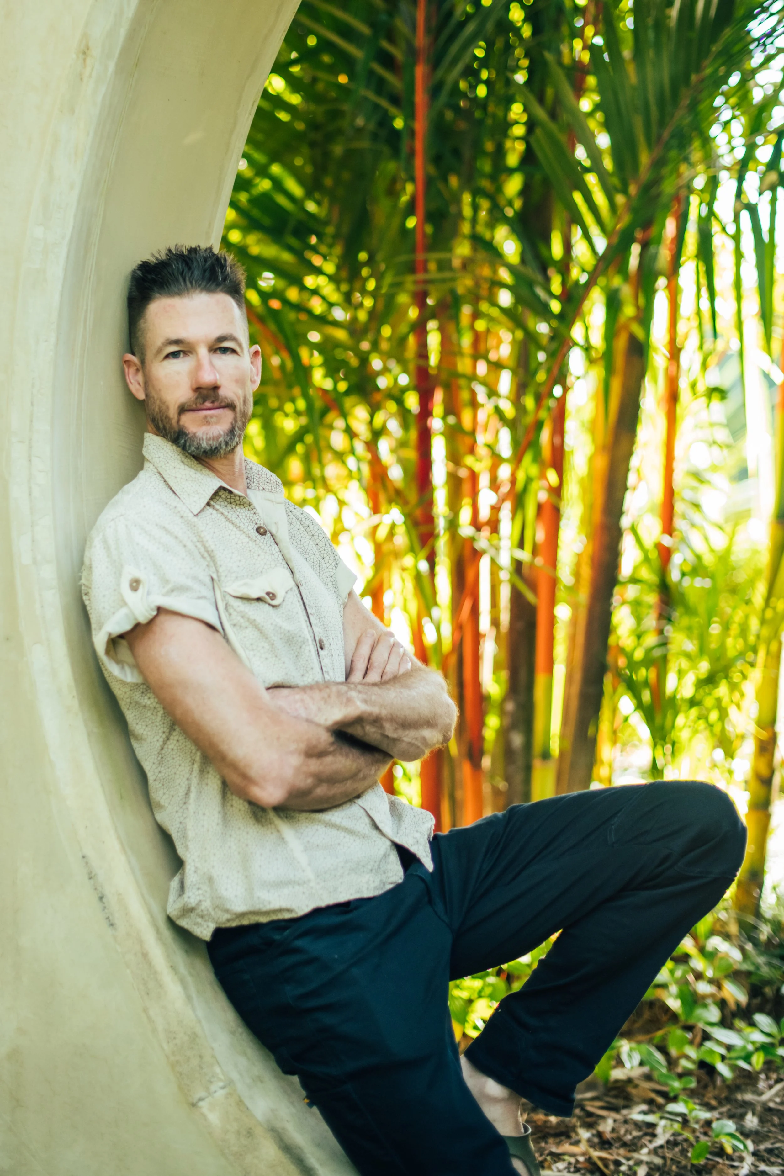 A man with dark hair and a beard, wearing a beige short-sleeve button-up shirt and dark pants, is sitting outdoors against a curved beige structure, with arms crossed and a neutral expression. He is surrounded by green foliage and trees.