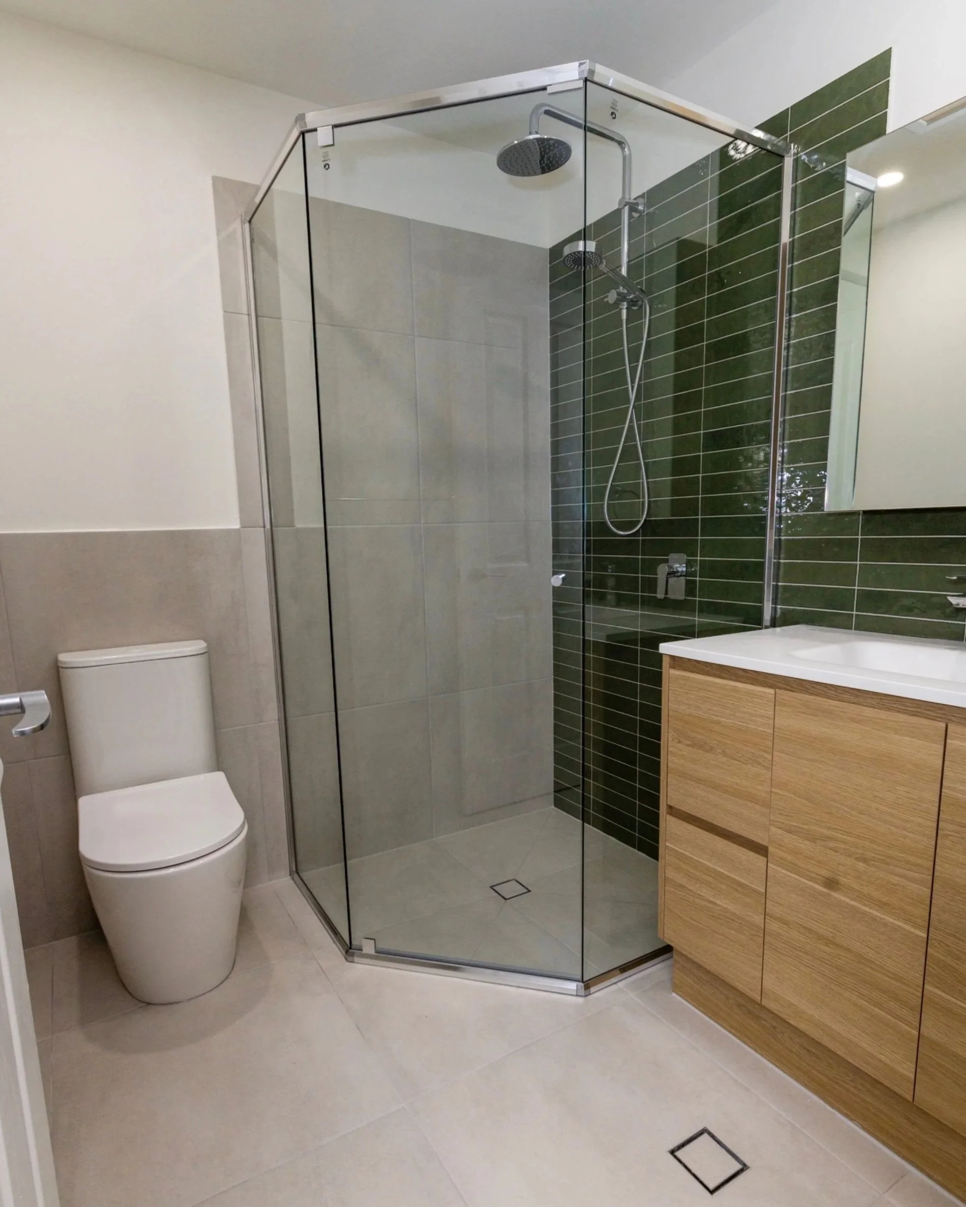 Modern bathroom with a toilet, a glass-enclosed shower with two showerheads, and a wooden vanity with a white sink, green tiled wall behind the shower, beige floor tiles, and a mirror above the sink.