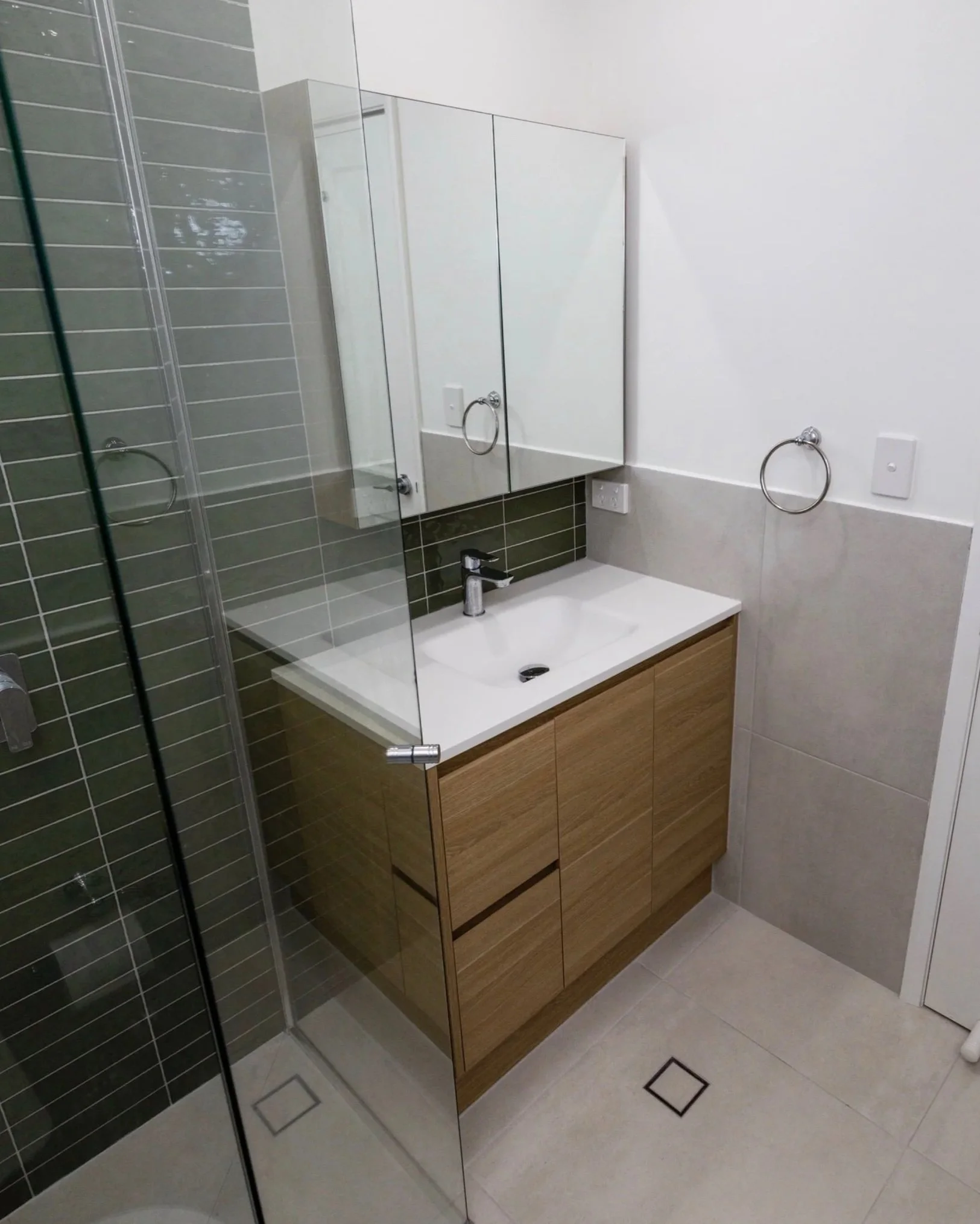 Modern bathroom with a glass-enclosed shower, a white vanity with a wooden cabinet, a mirror cabinet, towel rings on white walls, and tiled walls in white and dark green.