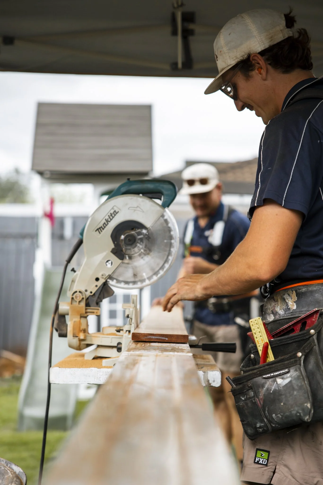 Two men working outdoors at a construction site, one cutting wood with a circular saw and the other smiling in the background.