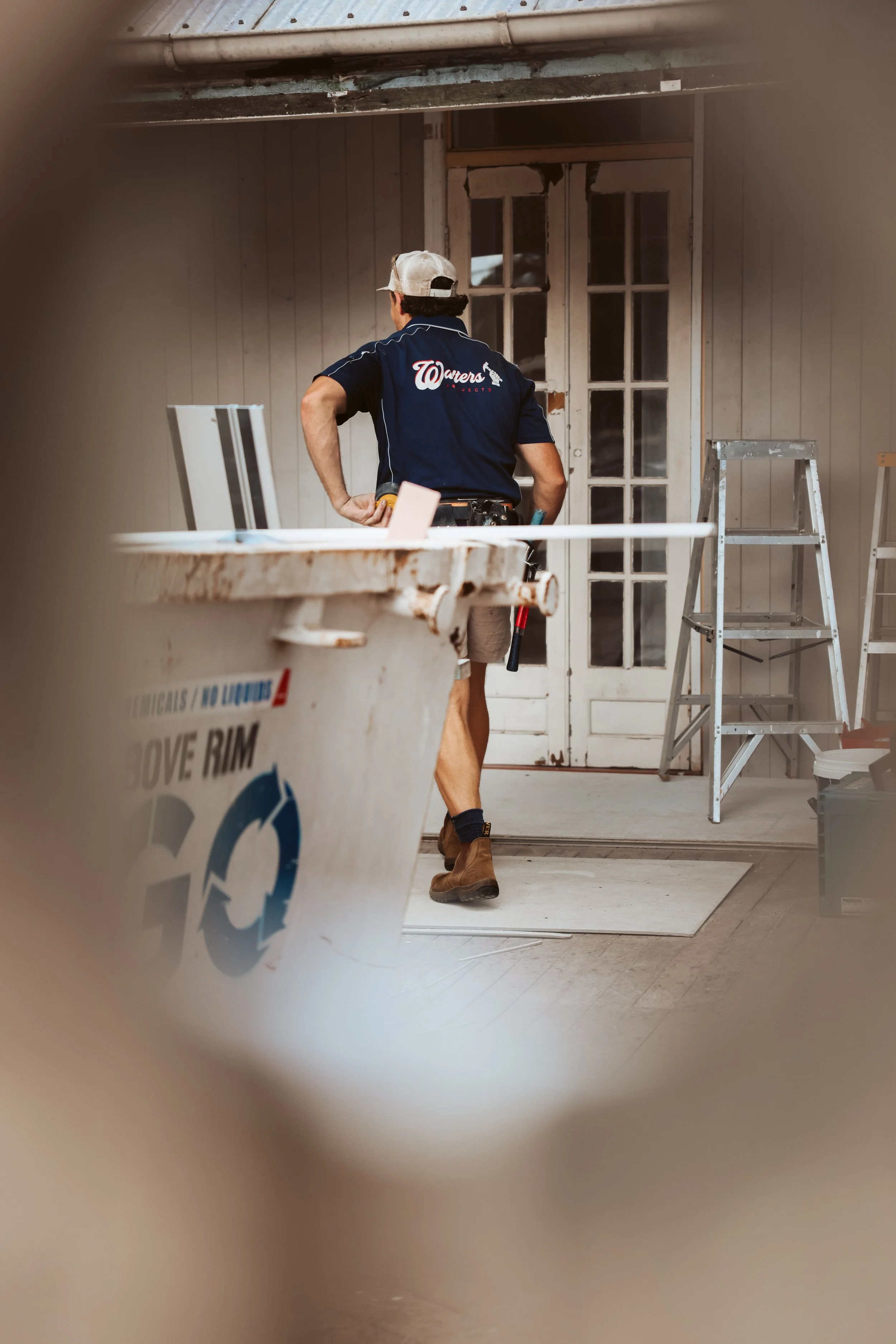 A man in work attire, standing in front of a door on a construction site, with ladders and construction tools around, viewed through a blurred framing.