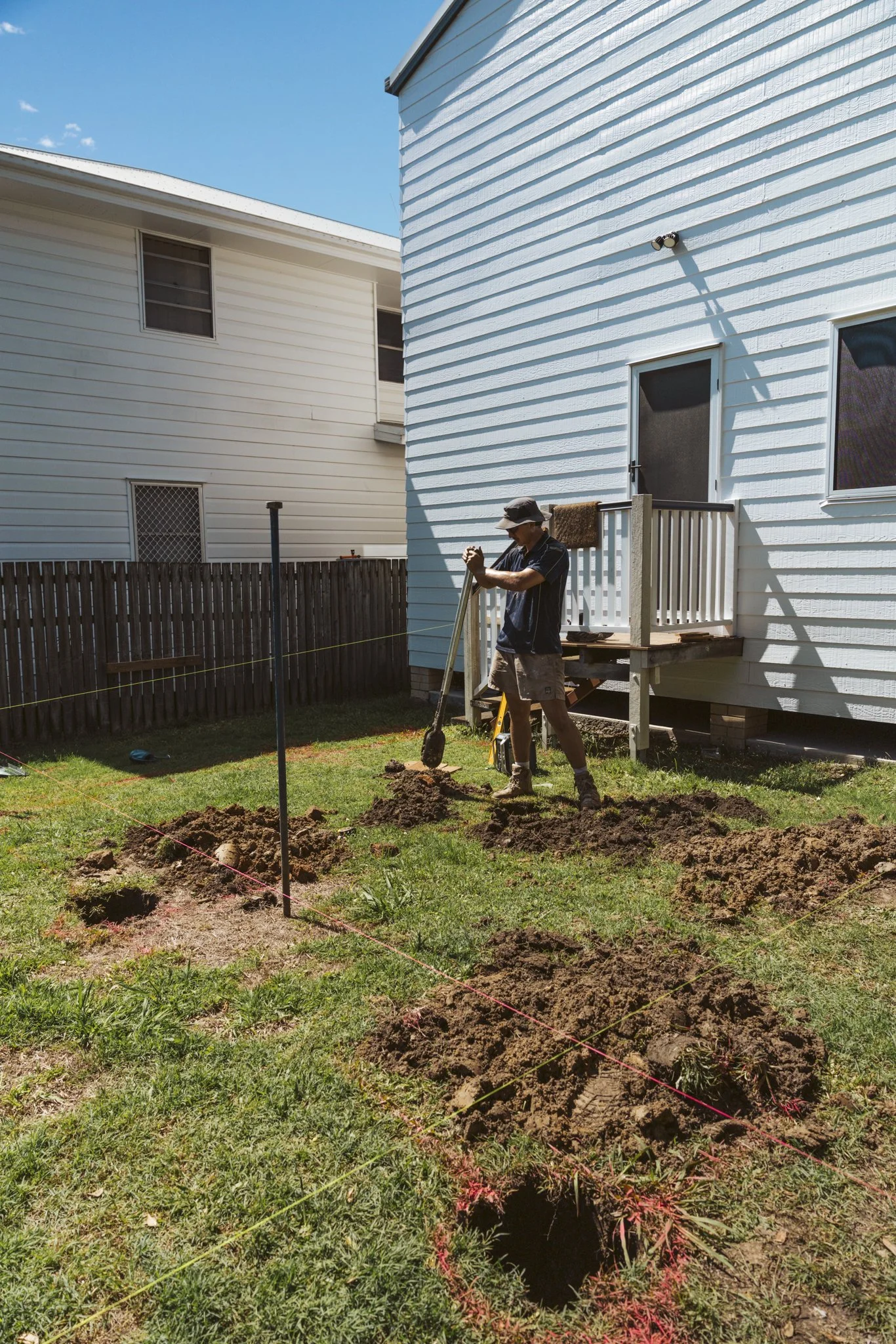 A man working on outdoor construction in a backyard during daytime, digging holes and using a tool, with a house and a wooden fence in the background.