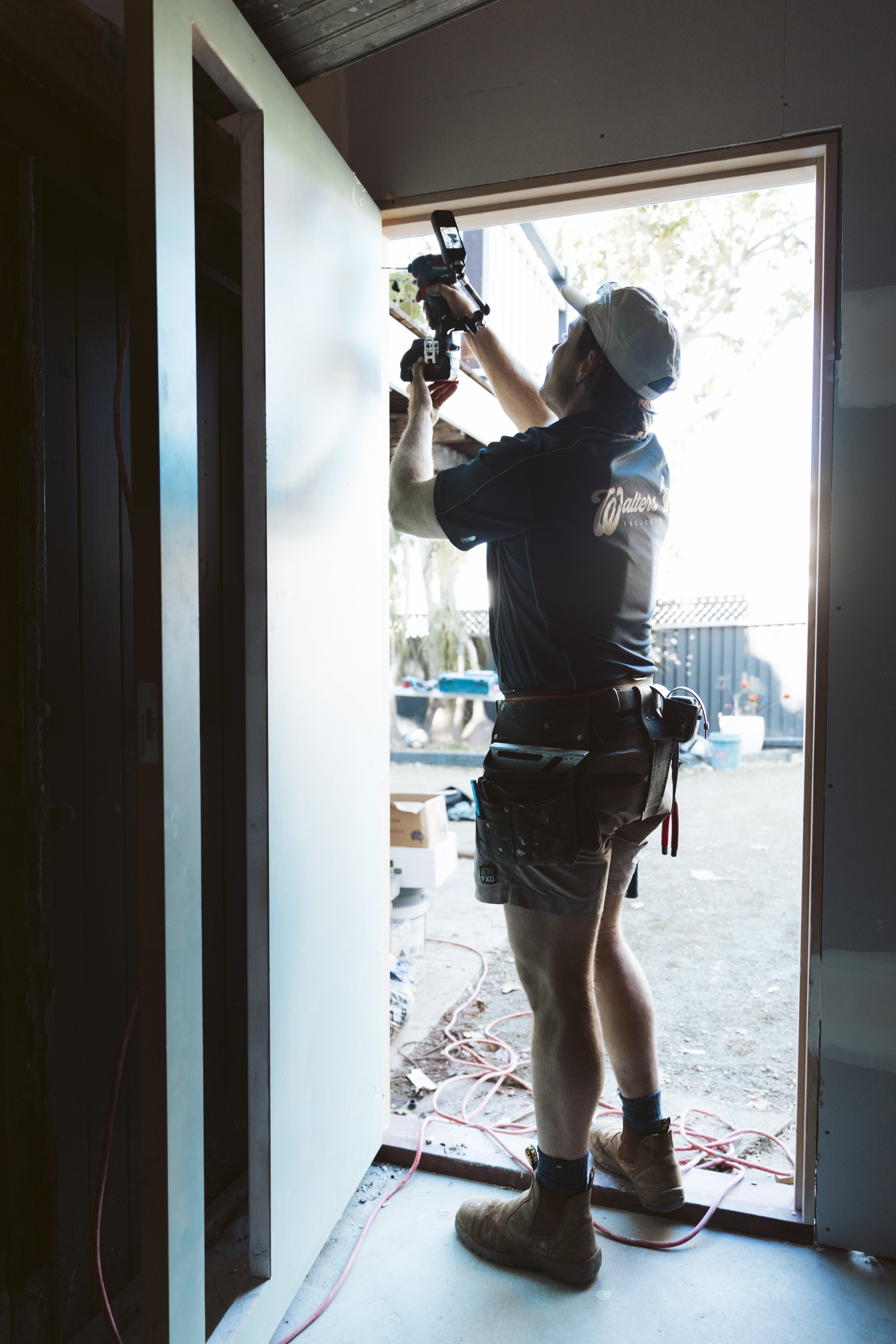 A construction worker installing or adjusting a door frame with a power tool outside a building. The worker is wearing a baseball cap, work boots, and a tool belt, standing in the doorway with sunlight and trees in the background.