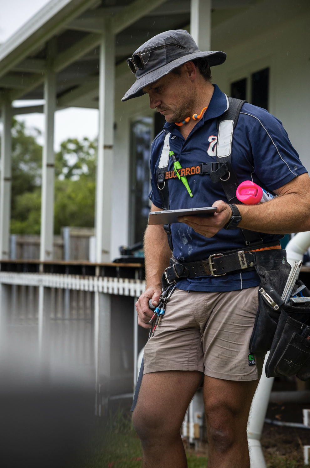 A man wearing a wide-brimmed hat, sunglasses, and outdoor gear, standing outside a house, looking at a tablet.
