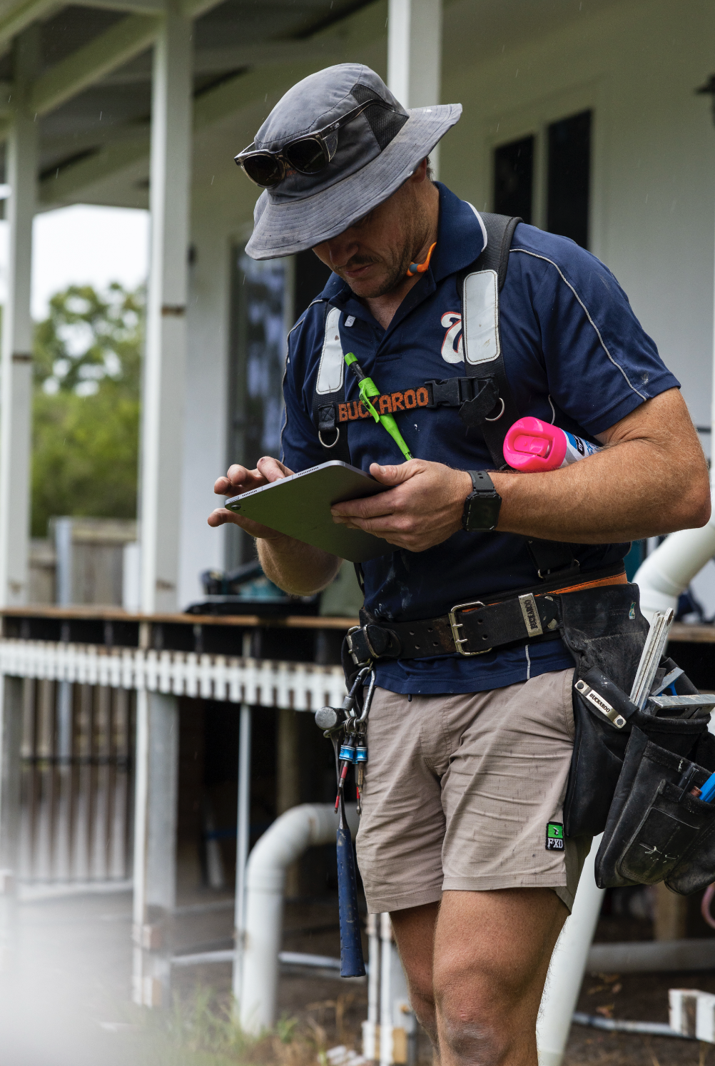 A man wearing a wide-brimmed hat, sunglasses, and outdoor gear looks at a tablet device, standing outside near a house with a porch.