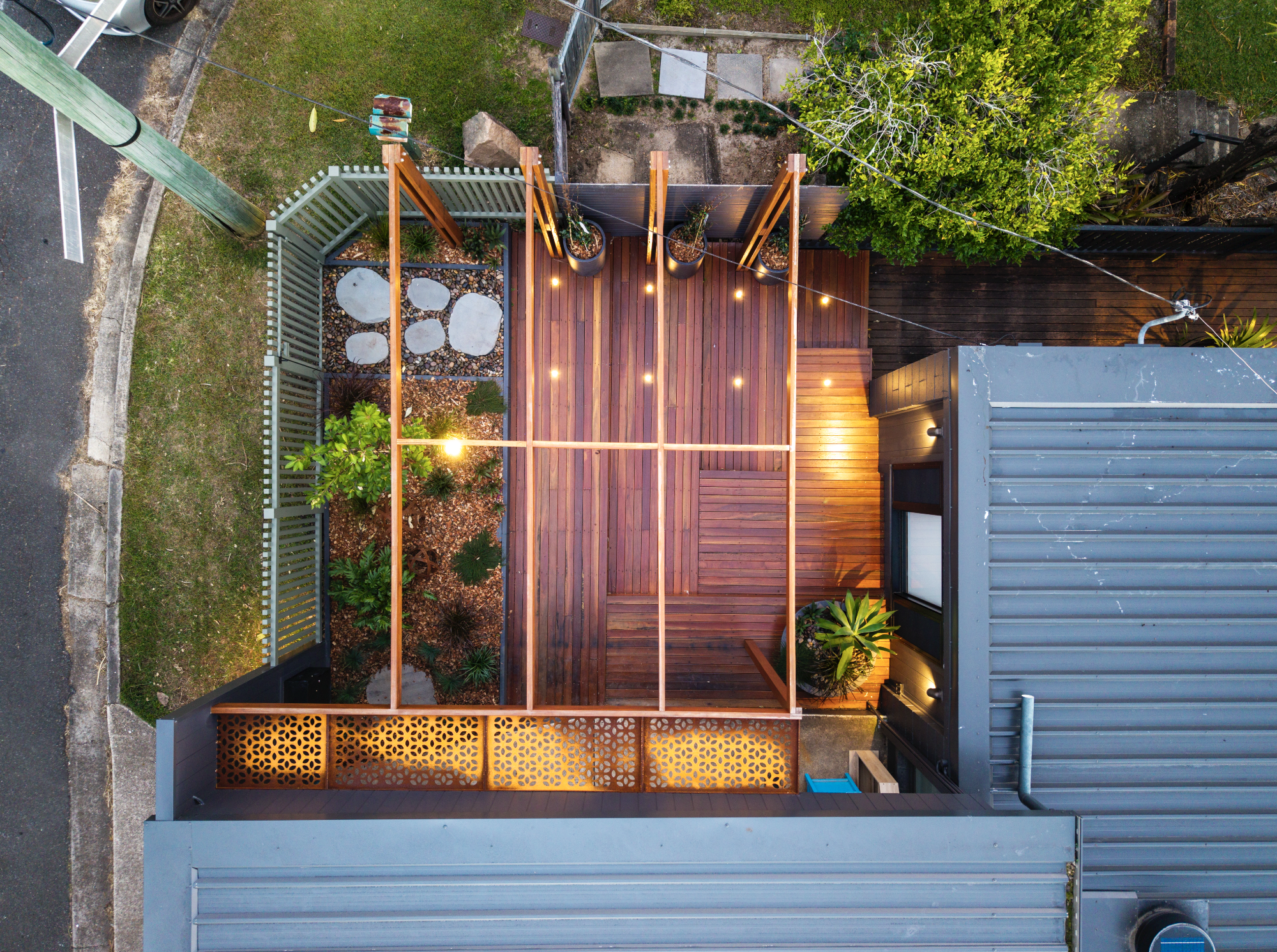 Top-down view of a backyard wooden deck area with string lights, potted plants, and a small garden with stones, surrounded by fencing, a gray house roof, and neighboring areas with grass and pavement.