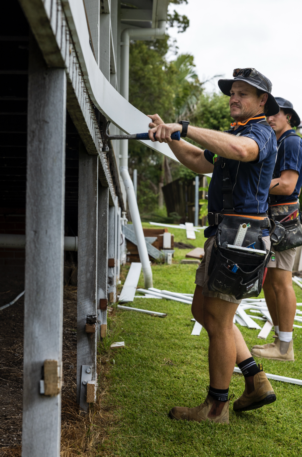 A man working outdoors on a construction project, wearing a hat and tool belt, holding a piece of white siding near a house's exterior wall, with another worker visible in the background.