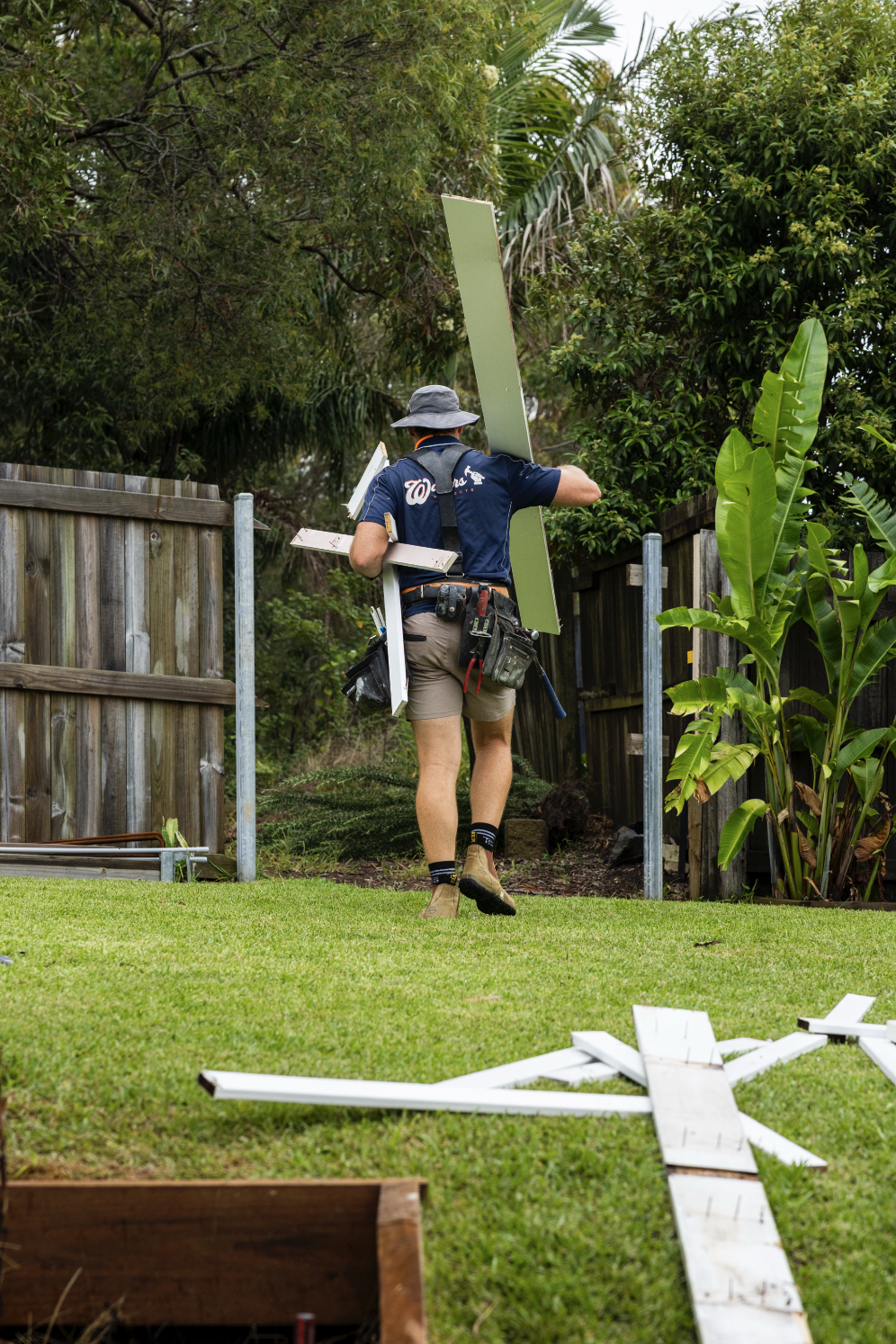 A man wearing a gray hat, navy shirt, and khaki shorts, carrying a large panel and other construction materials walks through a backyard with a wooden fence and green plants, assembling or installing a structure.