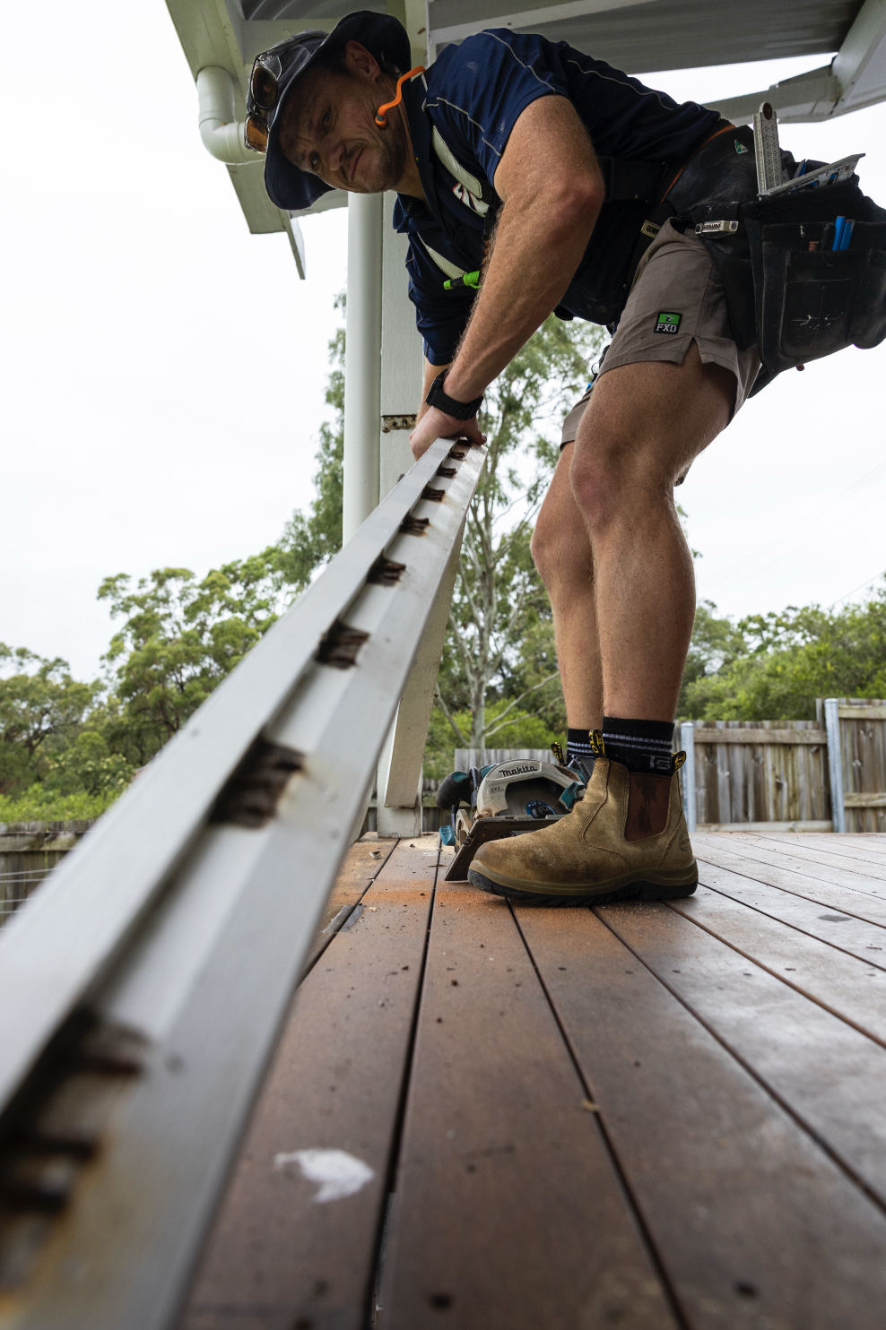 Man working on a wooden deck, using a measuring tape with a circular saw placed on the floor nearby, outdoor setting with trees and a fence.