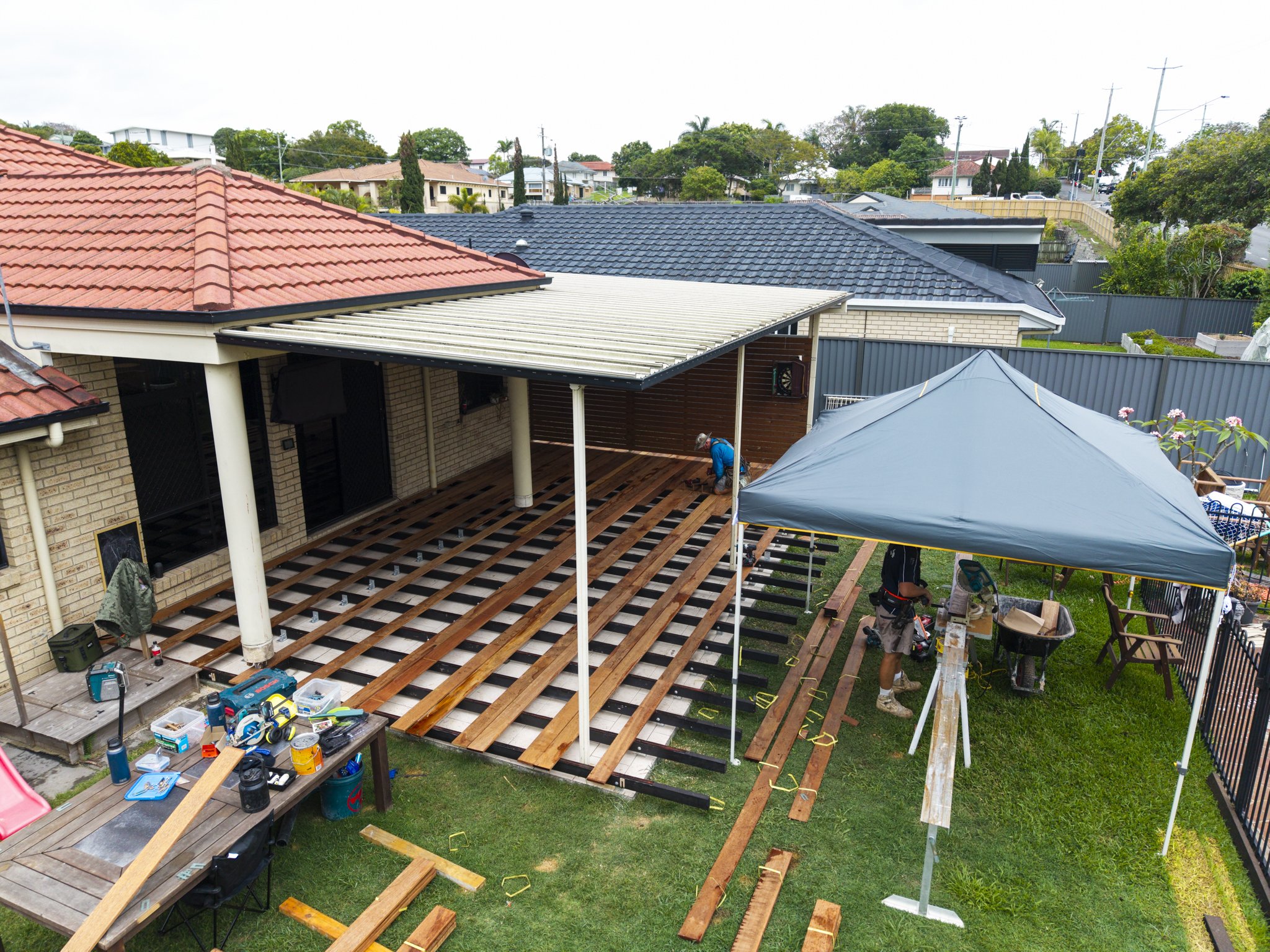 Backyard with house under renovation, workers installing a wooden deck, tools on table, and a blue tent
