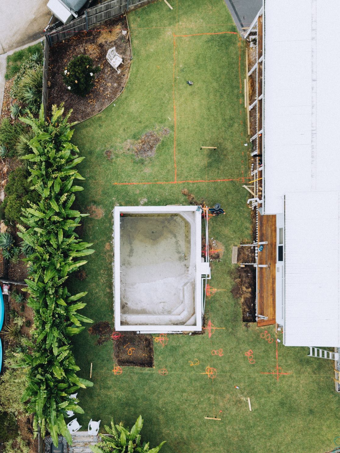 Aerial view of a backyard with an empty swimming pool under construction, a grassy lawn, a landscaped garden area, and a house with scaffolding.