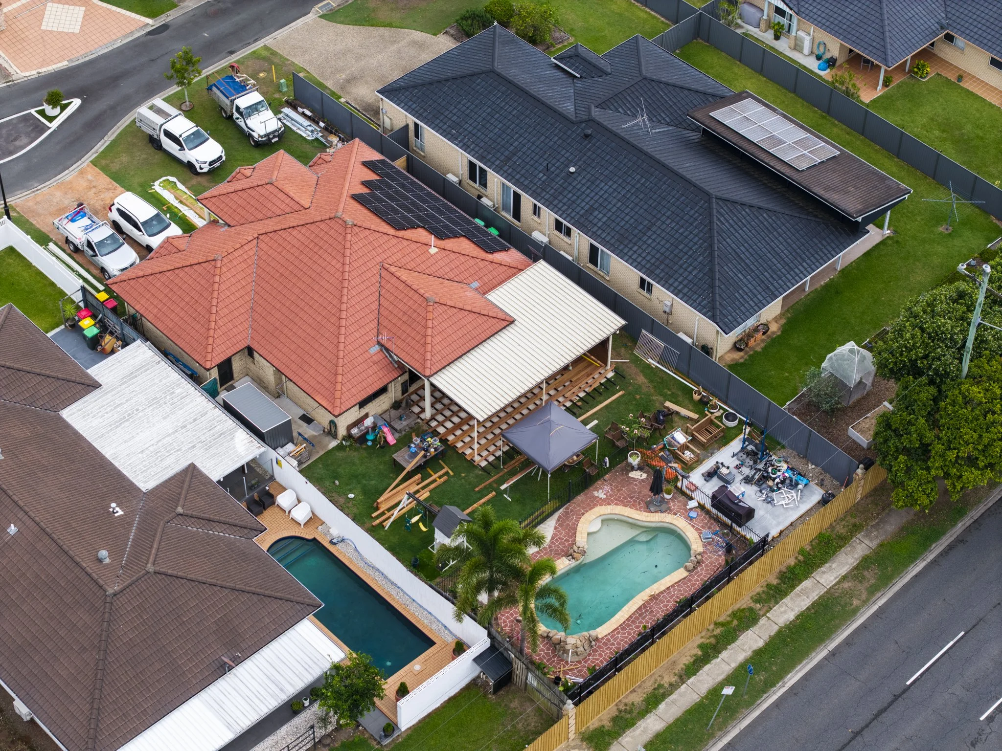 Aerial view of a suburban backyard with a pool, patio, and various outdoor furniture, surrounded by a fence and neighboring houses with different roof styles.