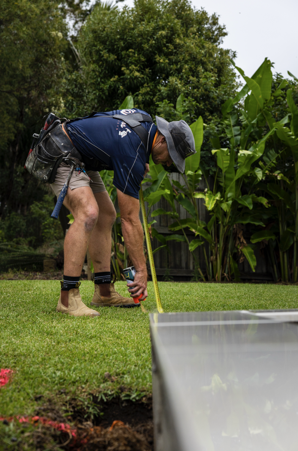 A man measuring the ground in a garden with a tape measure, wearing work boots, a hat, and a toolbox attached to his belt.