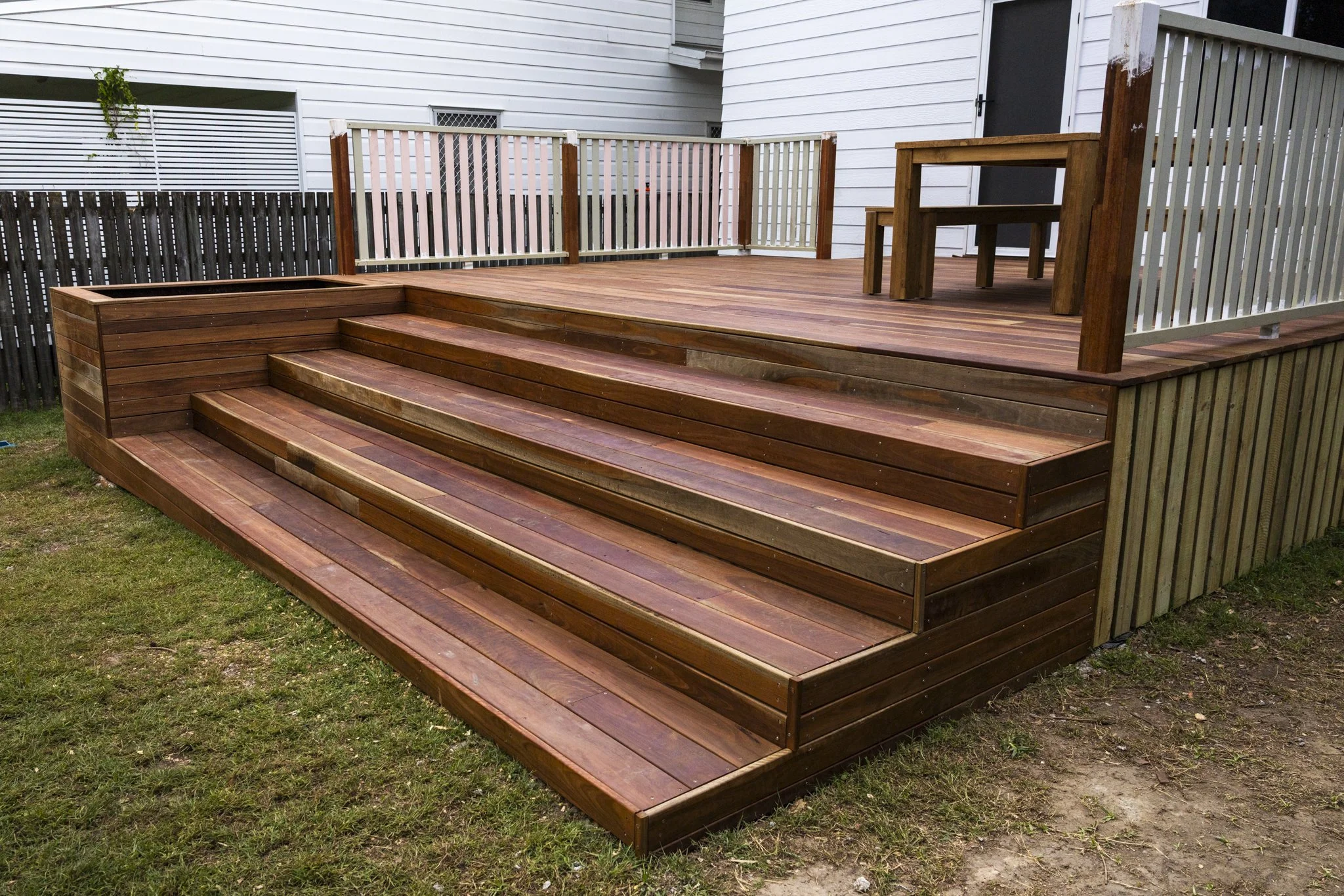 A wooden deck with stairs leading up to an elevated area with a picnic table and bench, surrounded by a white fence.